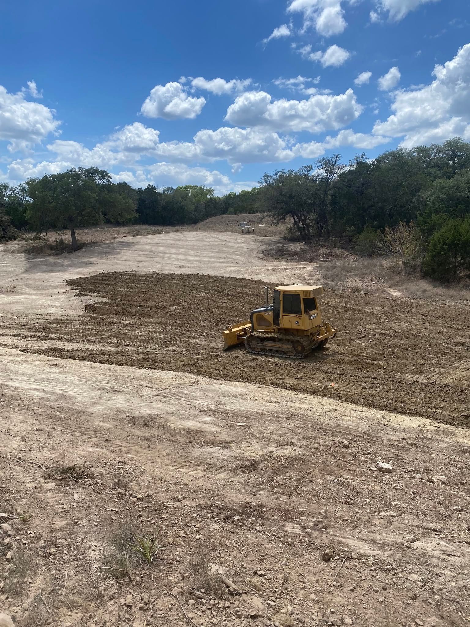 Yellow bulldozer leveling dirt in a dry clearing under a blue sky with clouds.