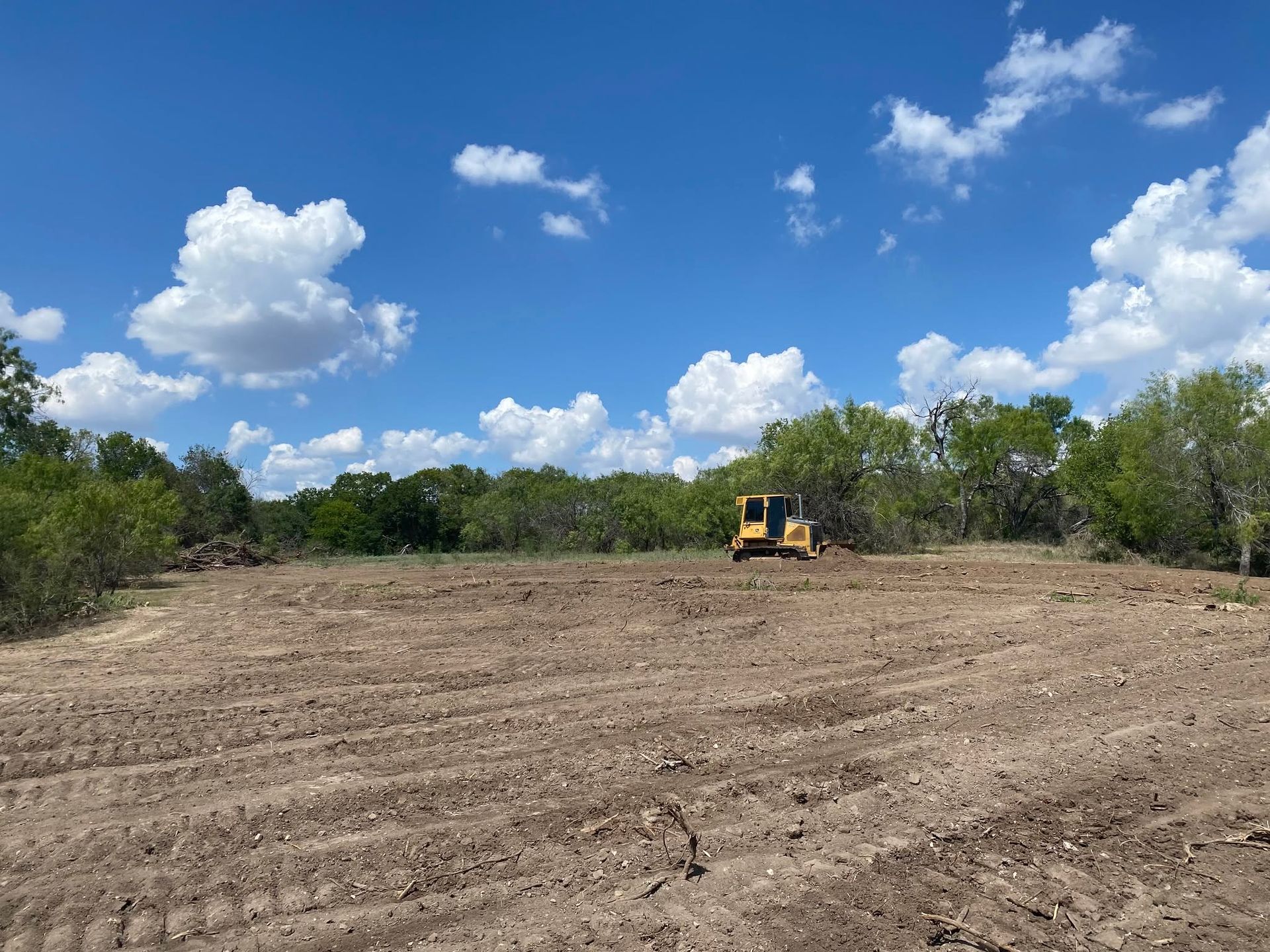 A yellow bulldozer levels a cleared dirt field under a bright blue sky with clouds.