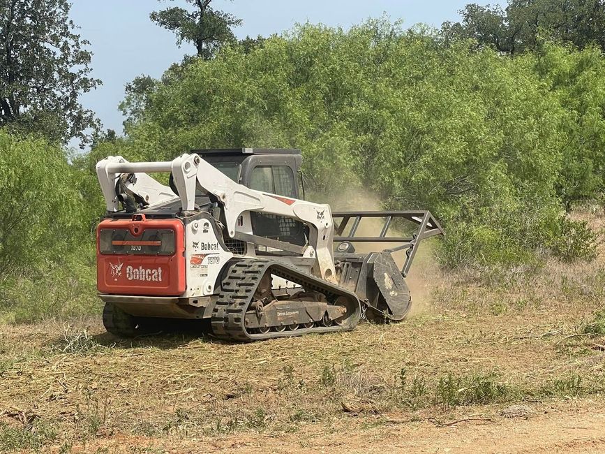 Small tracked skid steer clearing dry brush in a field with trees nearby