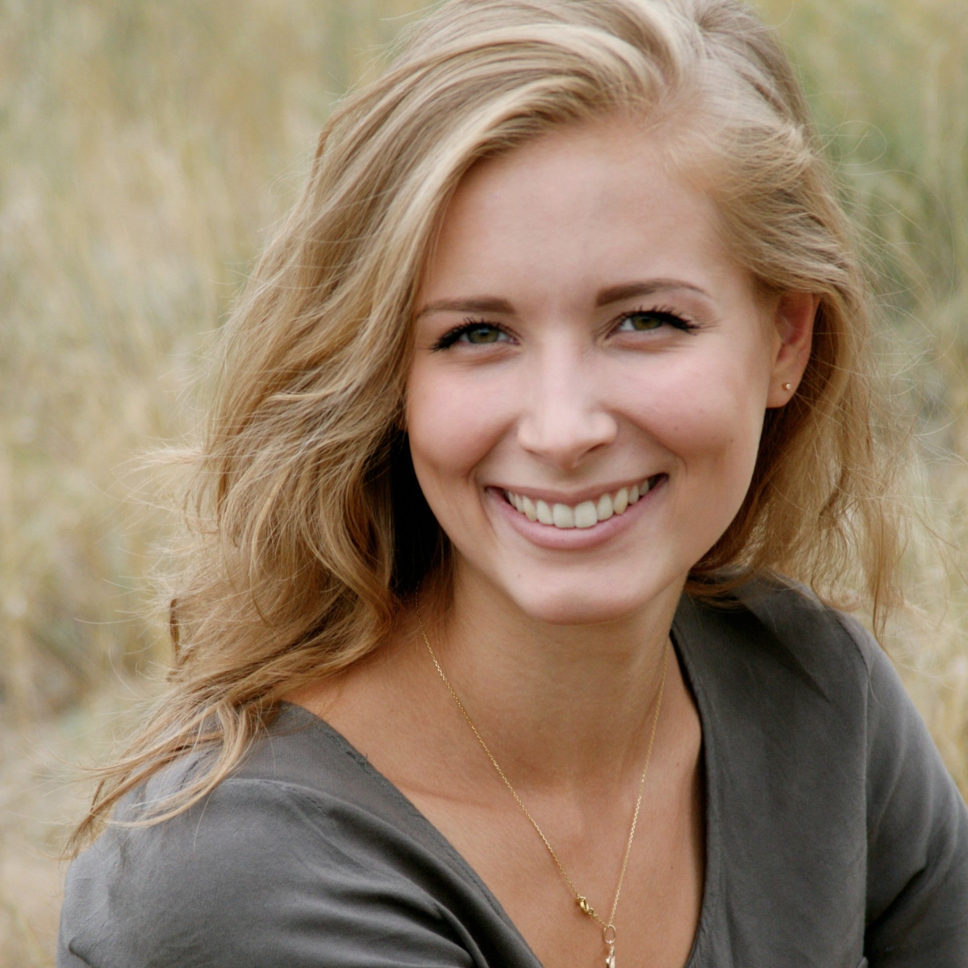 Smiling blonde woman in a gray top standing outdoors in tall grass