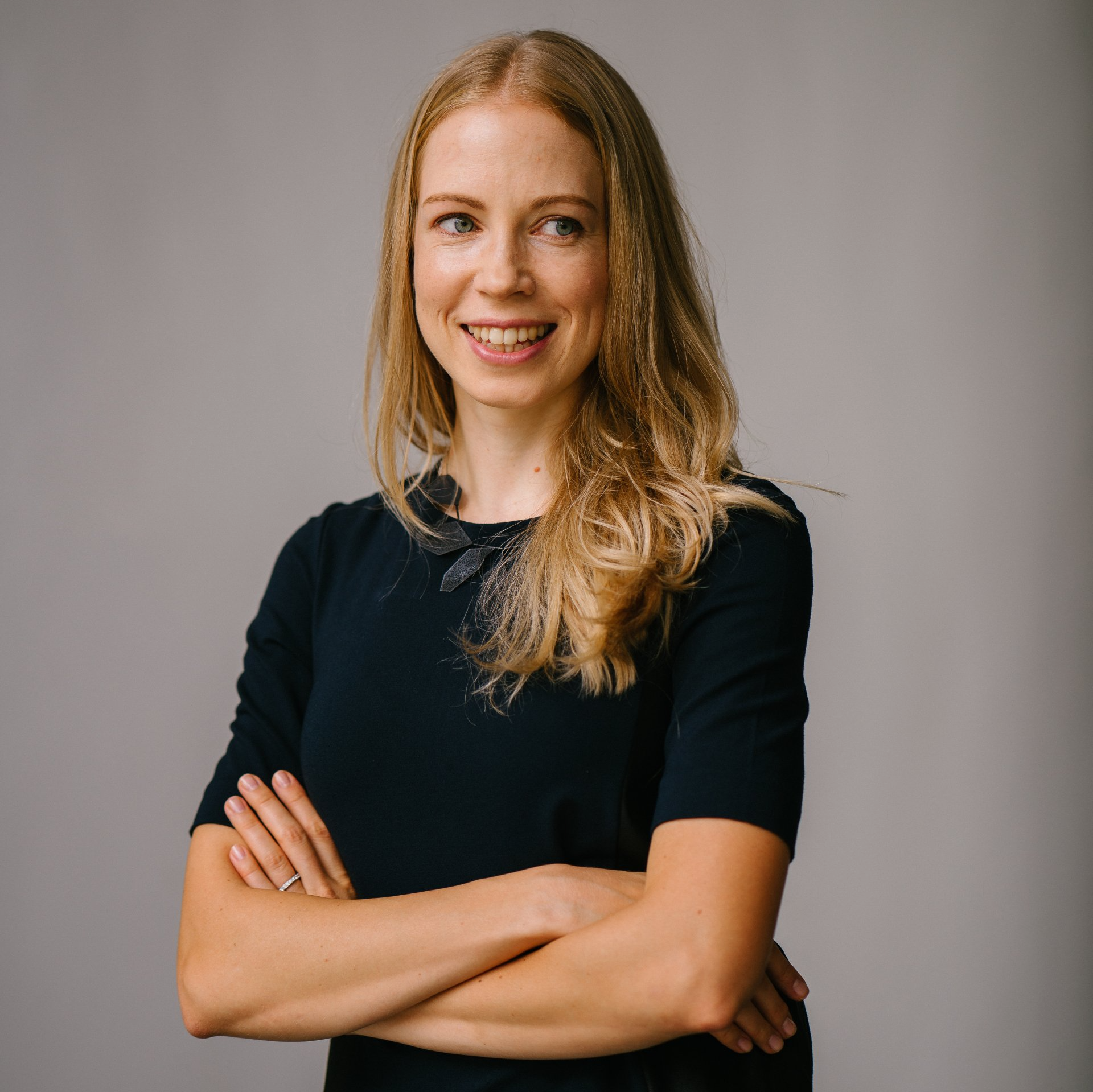 Smiling blonde person in a black shirt with arms crossed against a gray background