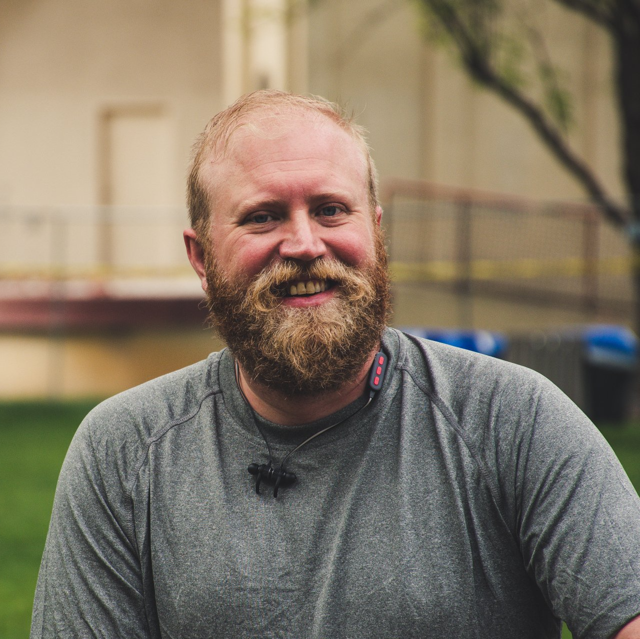 Smiling bearded man in a gray shirt outdoors, with a blurred building and trees behind him