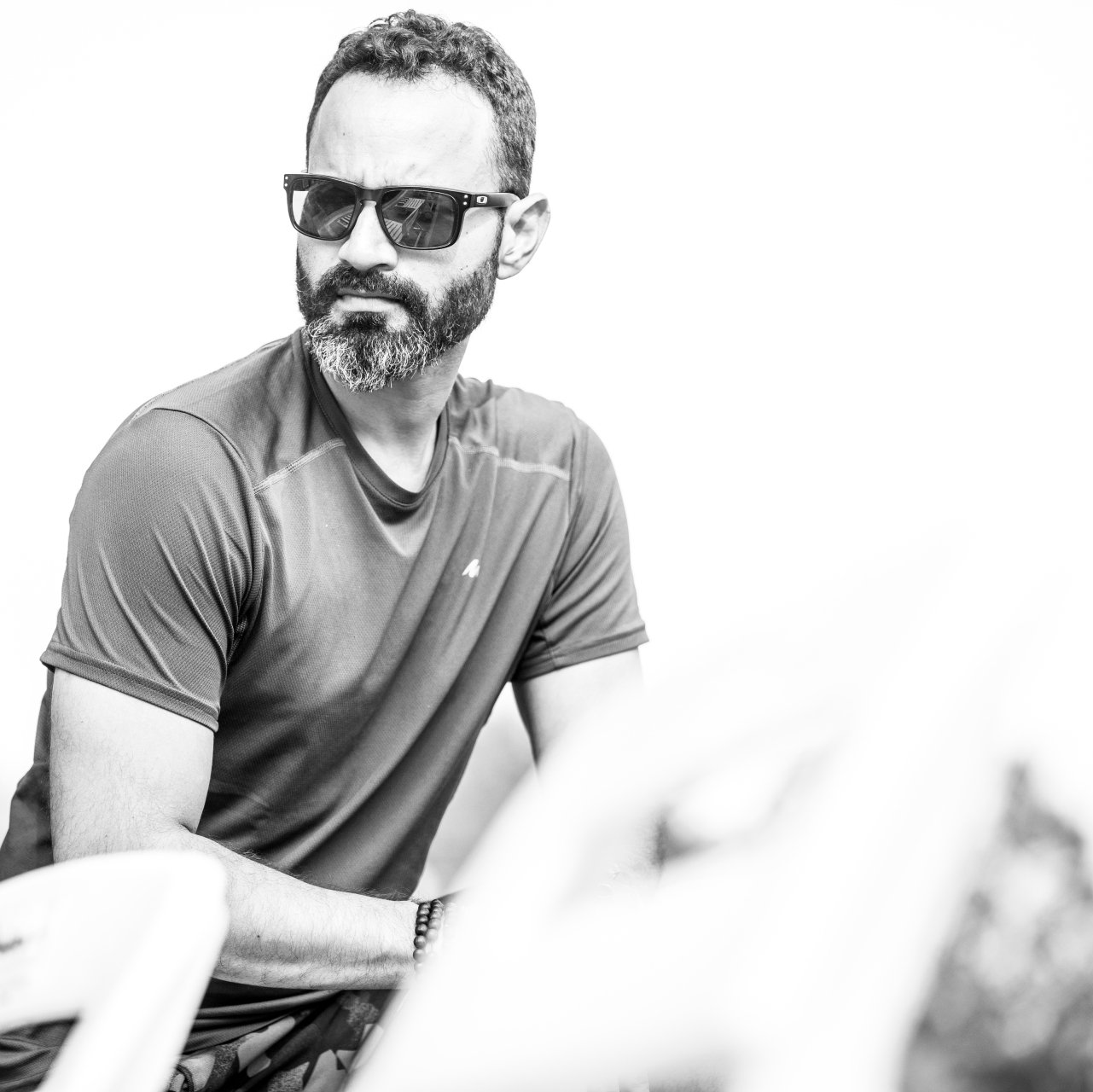 Man in sunglasses and a dark T-shirt sitting outdoors, looking to the side.