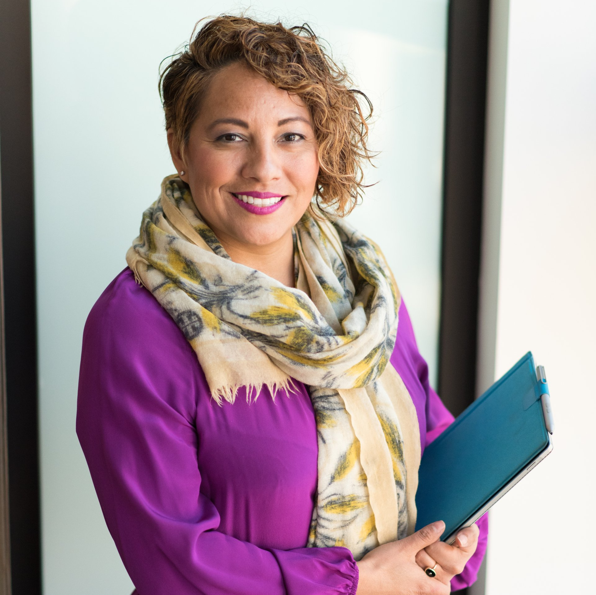 Smiling person in purple top and patterned scarf holding a blue folder against a light background