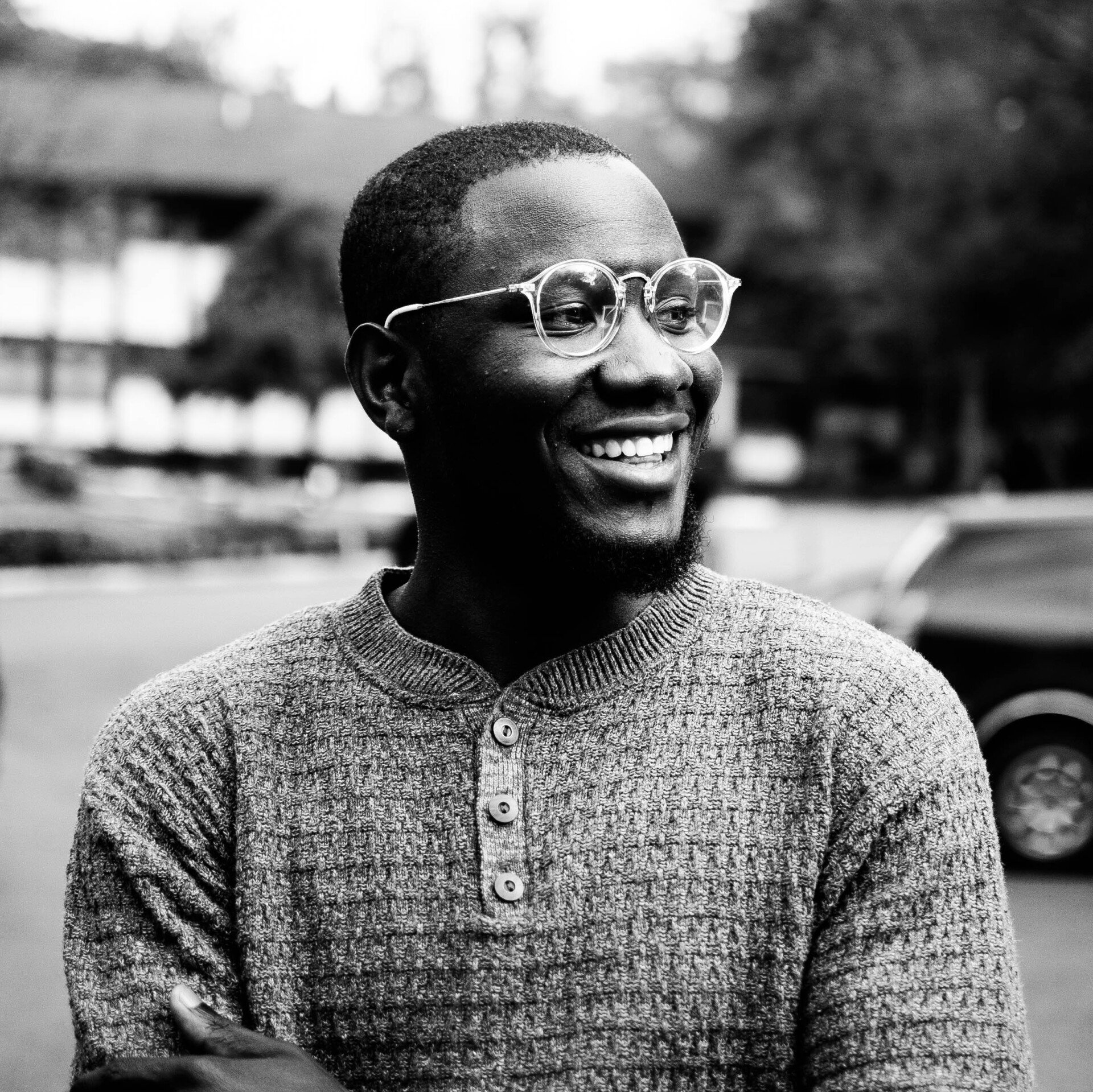 Smiling person in glasses and a textured sweater outdoors, with cars and buildings blurred in the background