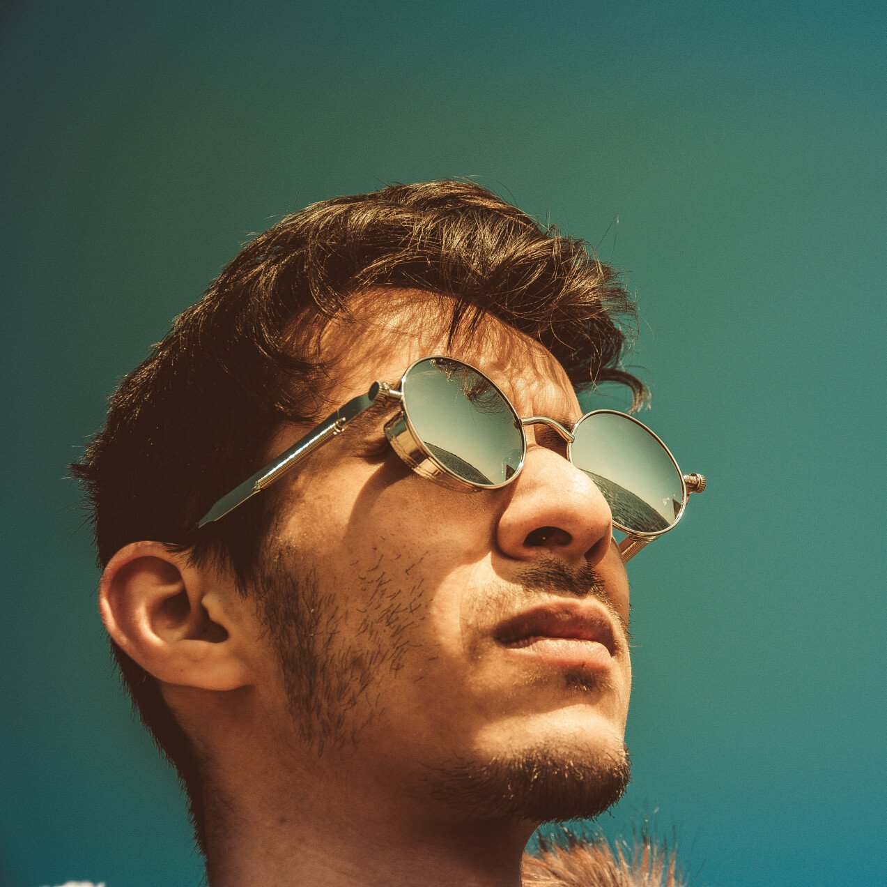 Close-up of a man wearing reflective sunglasses against a teal background, looking upward.