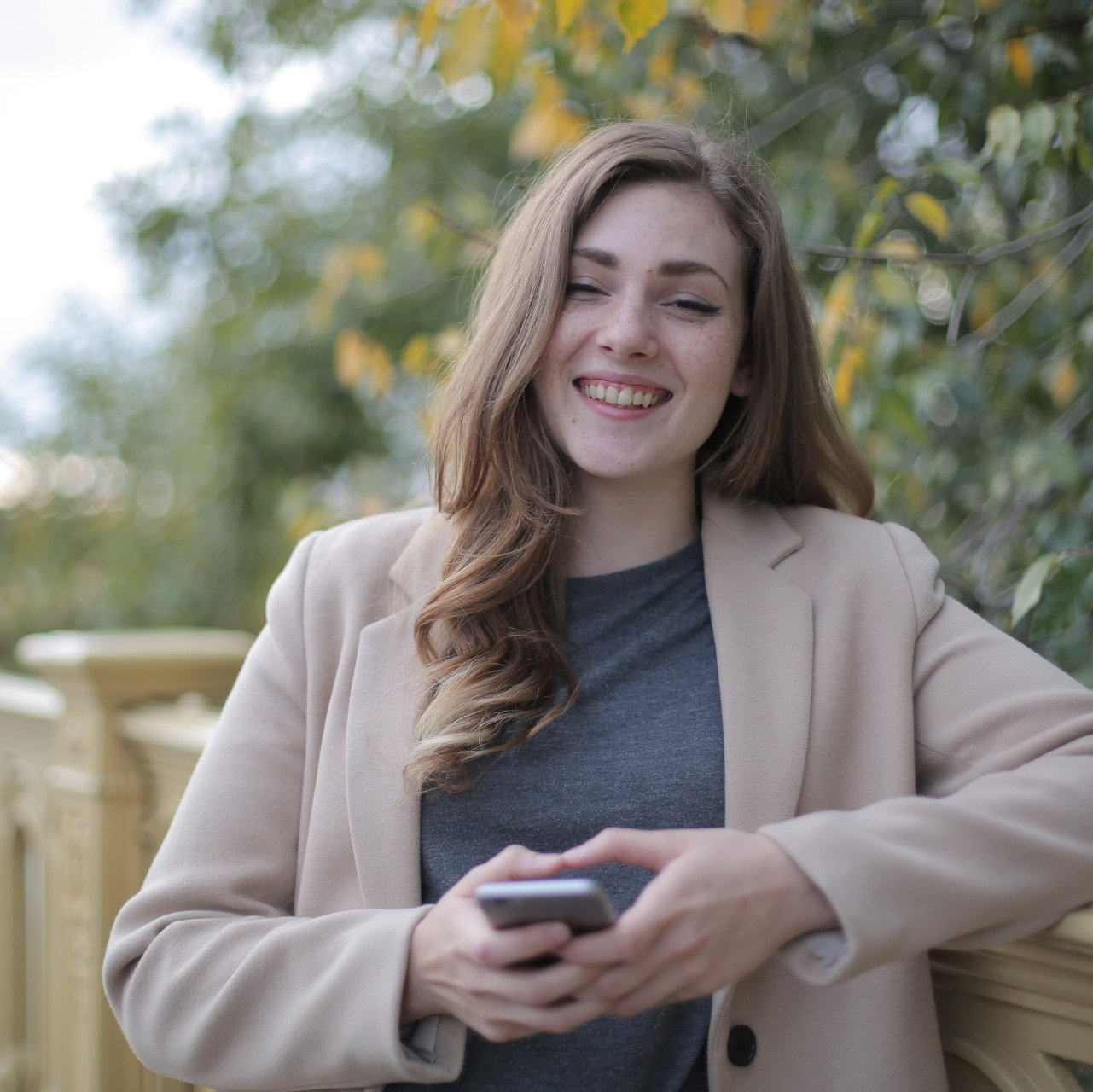 Woman smiling on a wooden deck, holding a phone, with trees blurred in the background