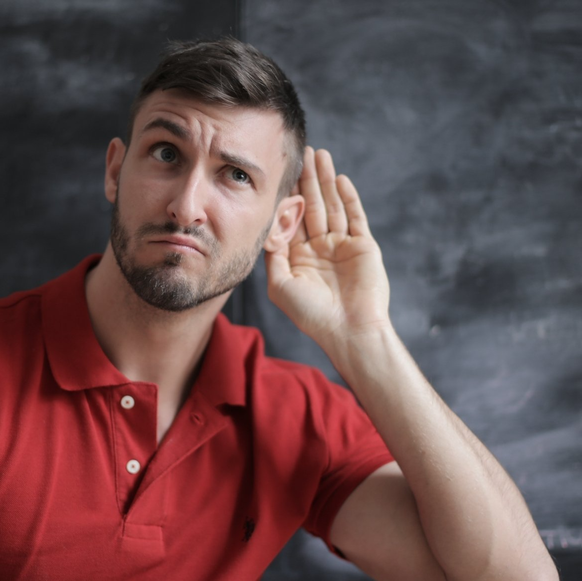 Man in a red shirt cupping his ear, listening with a puzzled expression against a gray background