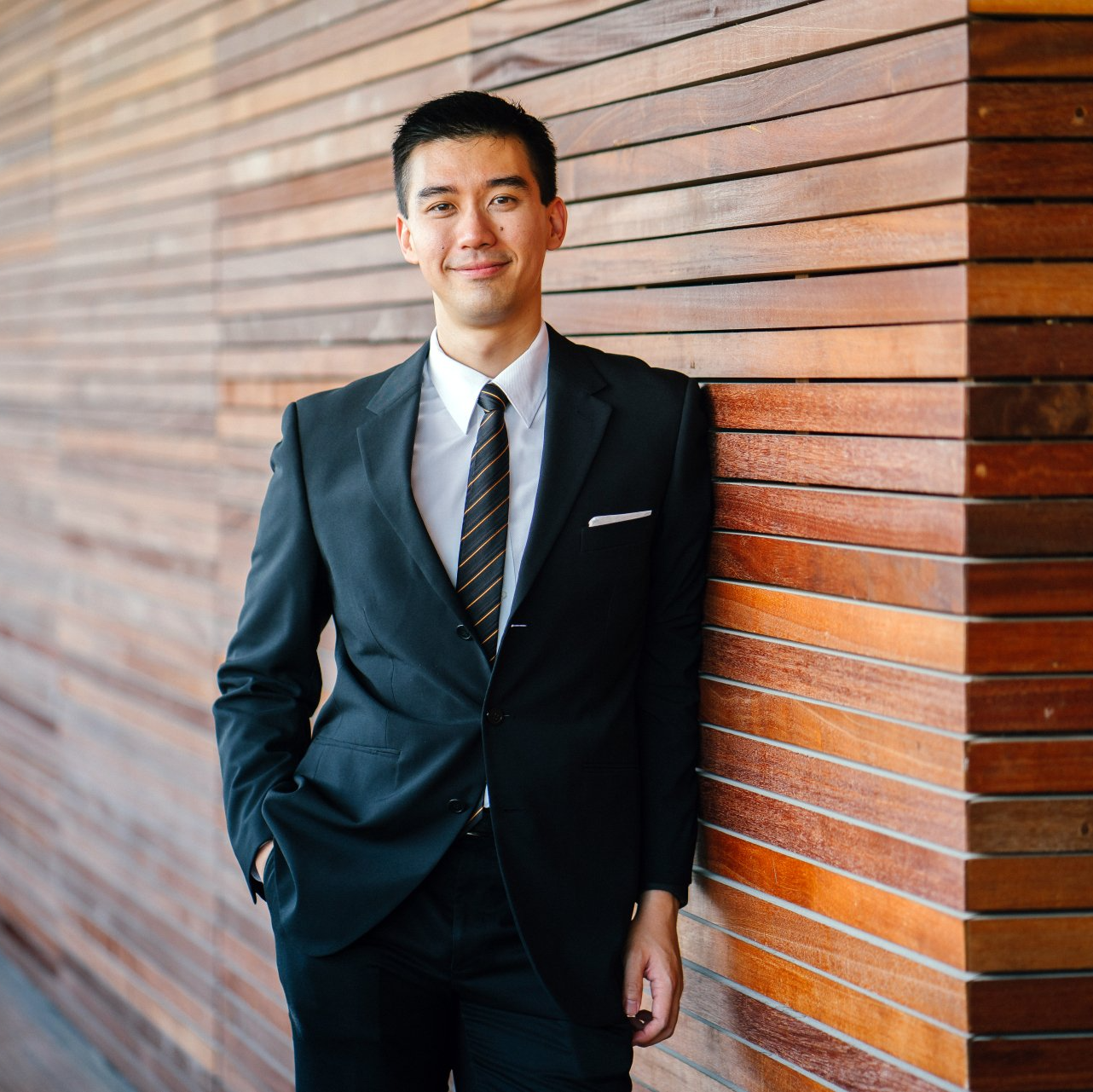 Man in a black suit leaning against a wooden wall, smiling gently.