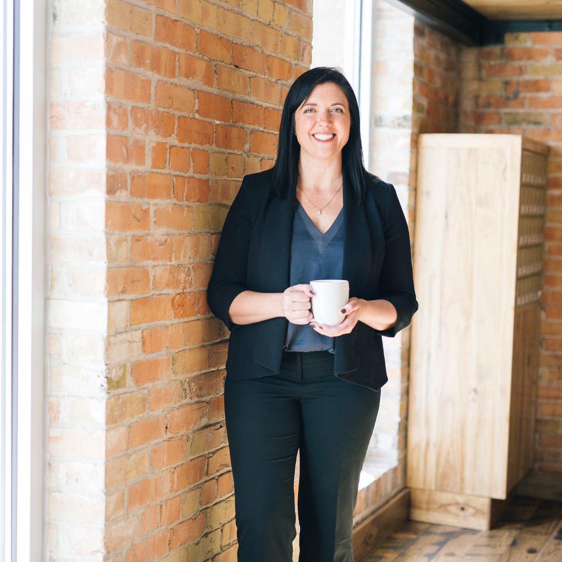 Woman in black suit holding a mug, standing by a brick wall in a bright hallway.
