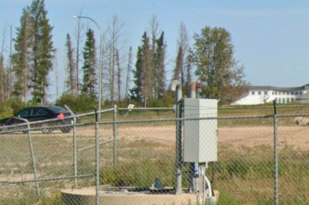 A gray electrical box and pipes enclosed by a chain-link fence, with a car and buildings in the background.