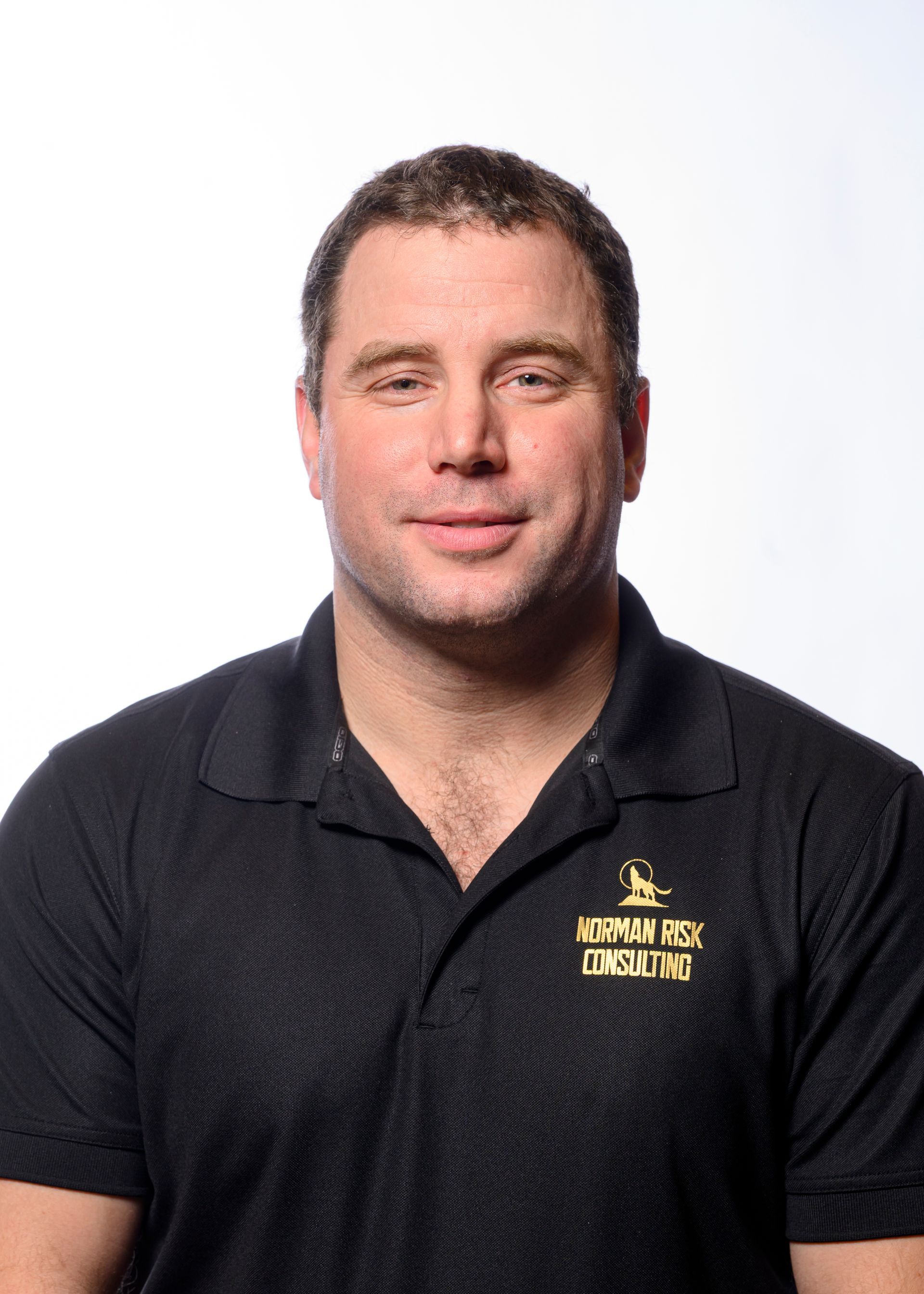 Man in black polo shirt, smiling, with logo on shirt. White background.