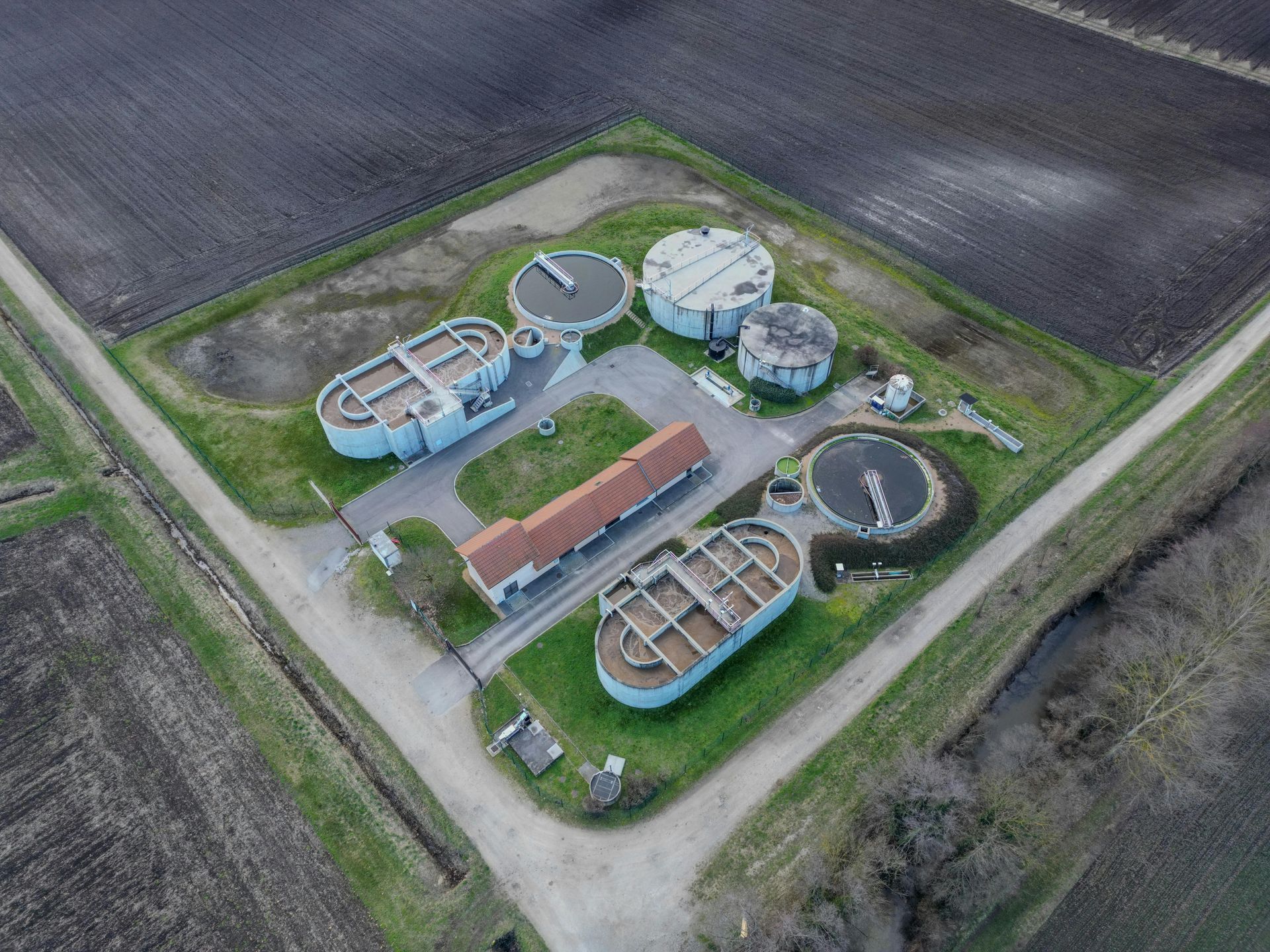 Aerial view of a wastewater treatment plant surrounded by farmland, with multiple circular and rectangular tanks.