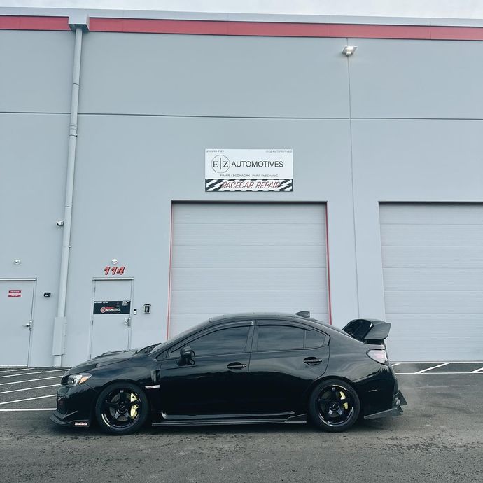 Black sports car parked beside a gray industrial building with a roll-up garage door.