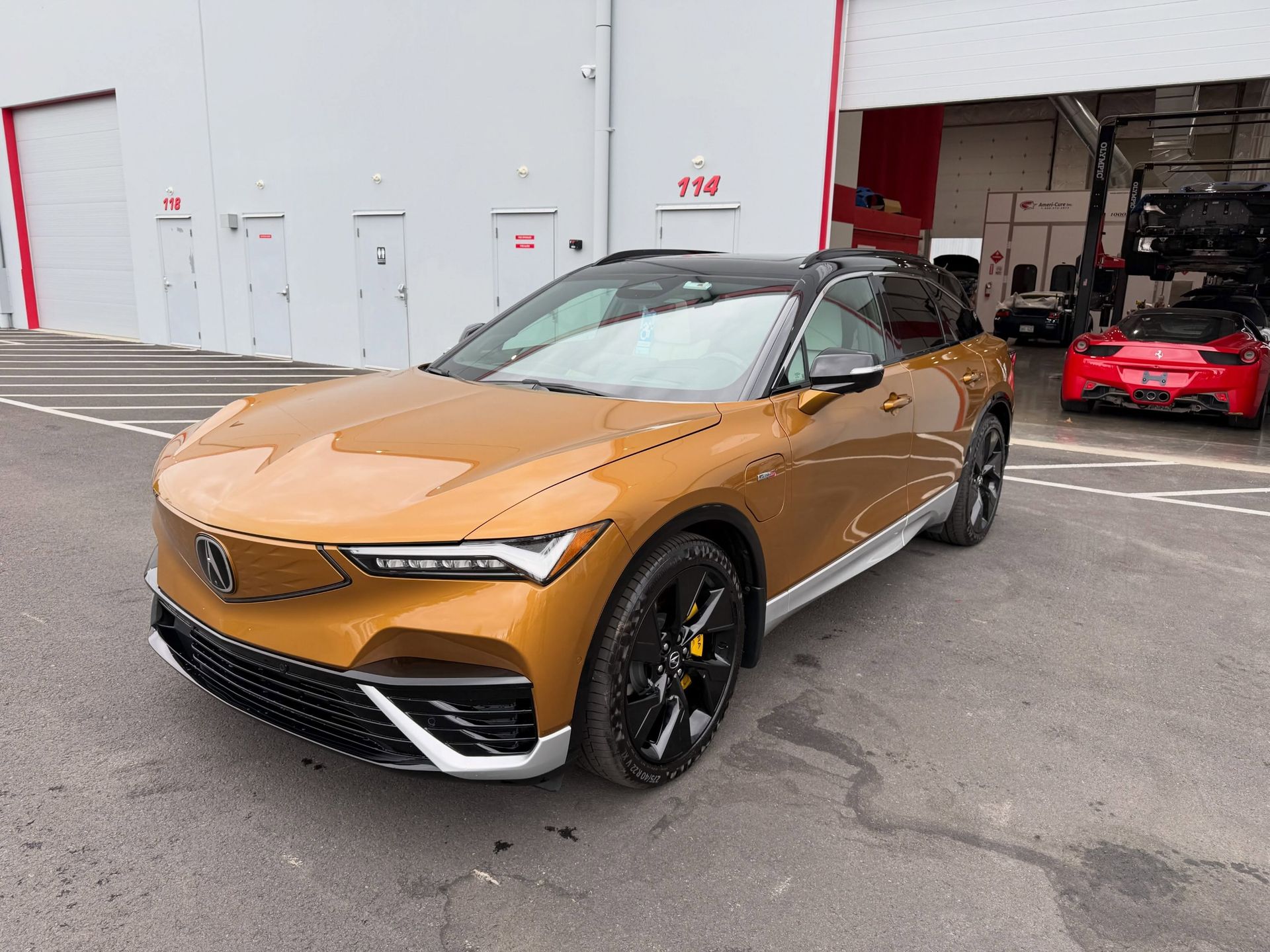 Copper Acura SUV parked outside a warehouse, with a red sports car in the background.