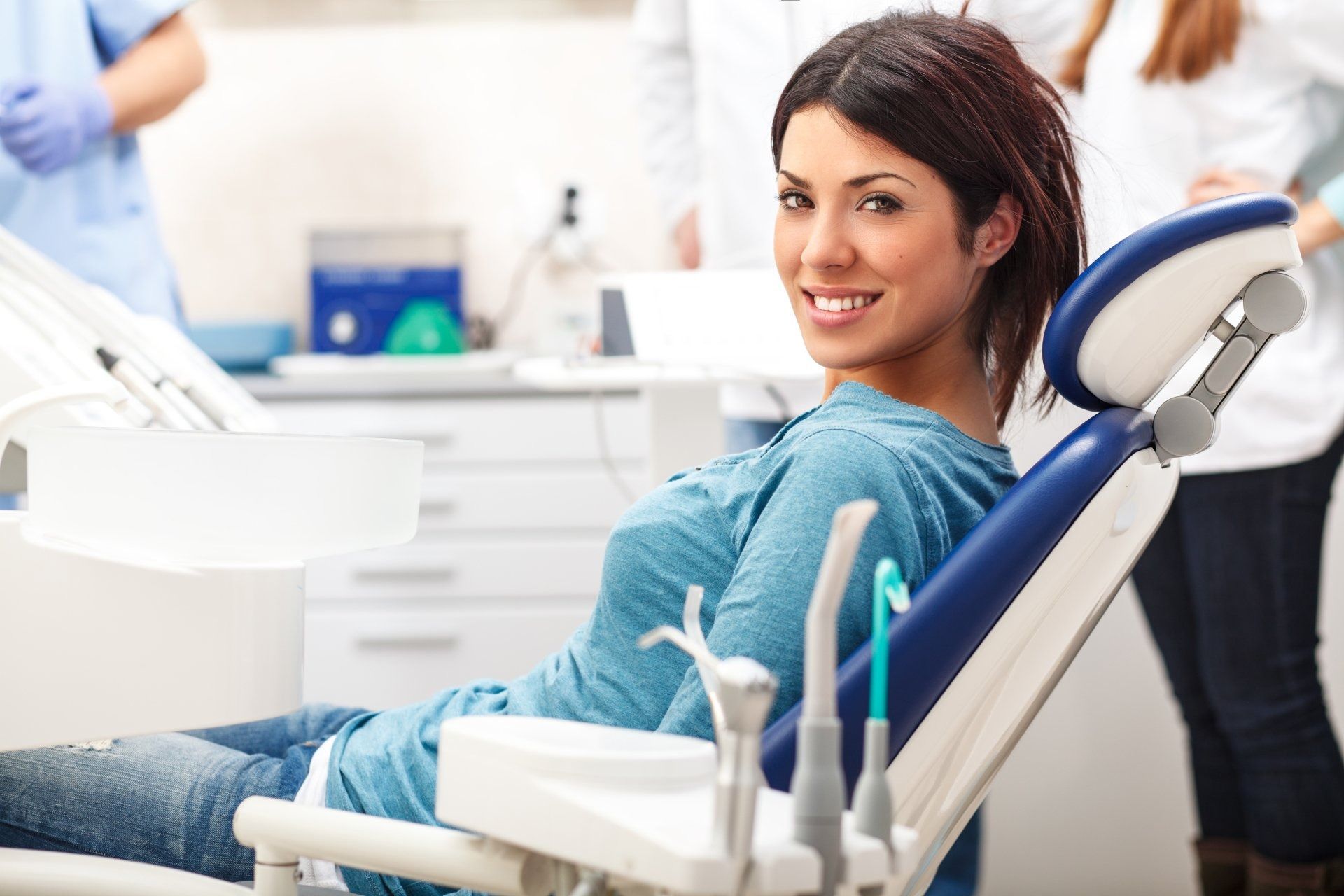 Smiling patient reclining in a blue dental chair during a dental exam, with tools and staff in a bright clinic