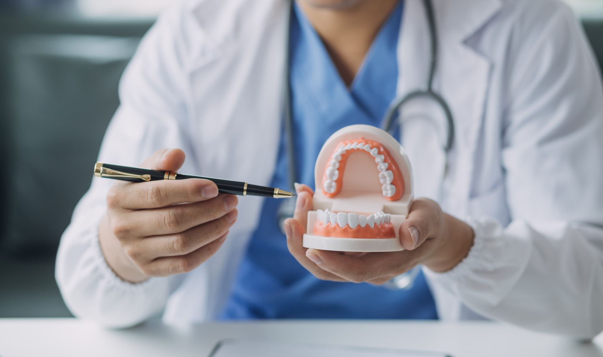Dentist holding a dental model and pointing at teeth with a pen, wearing blue scrubs and a white coat.