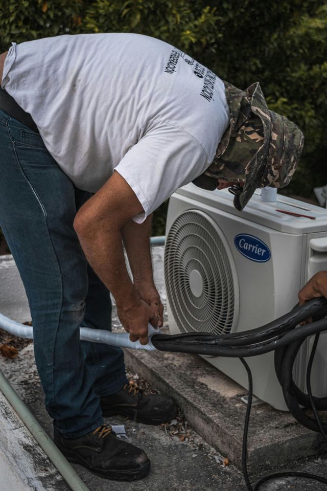Man in white shirt and camo hat working on an outdoor air conditioning unit on a rooftop.