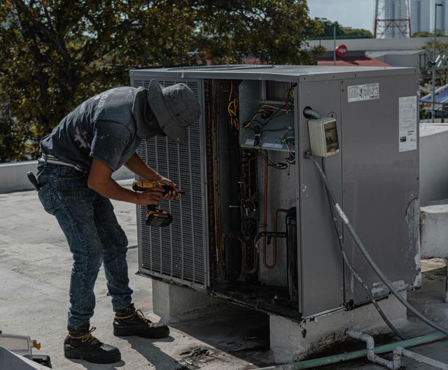 HVAC technician in jeans and hat repairs air conditioner unit on rooftop, using a power drill.