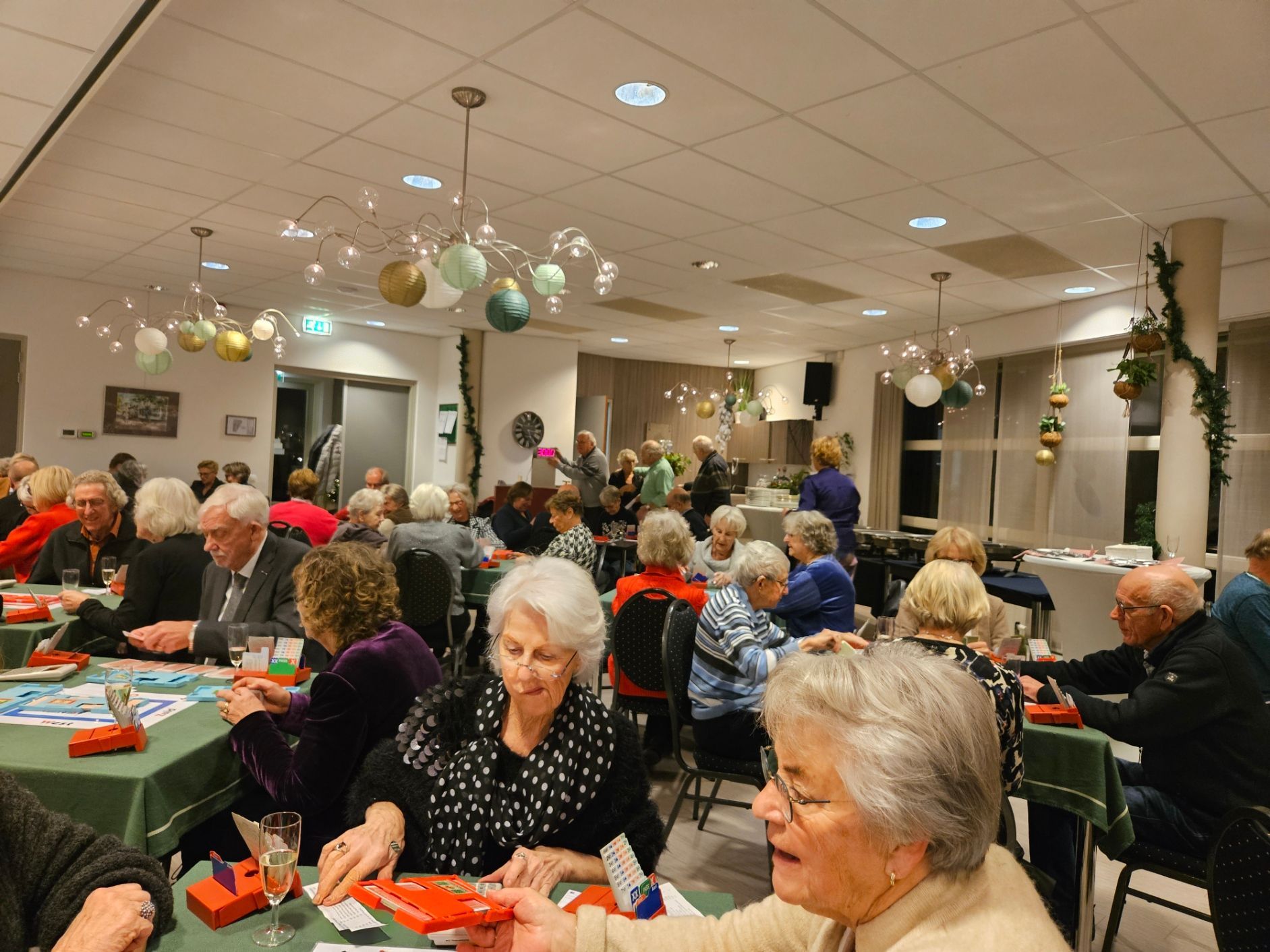 Mensen zitten aan tafels en spelen een spel in een versierde kamer met plafondlampen.