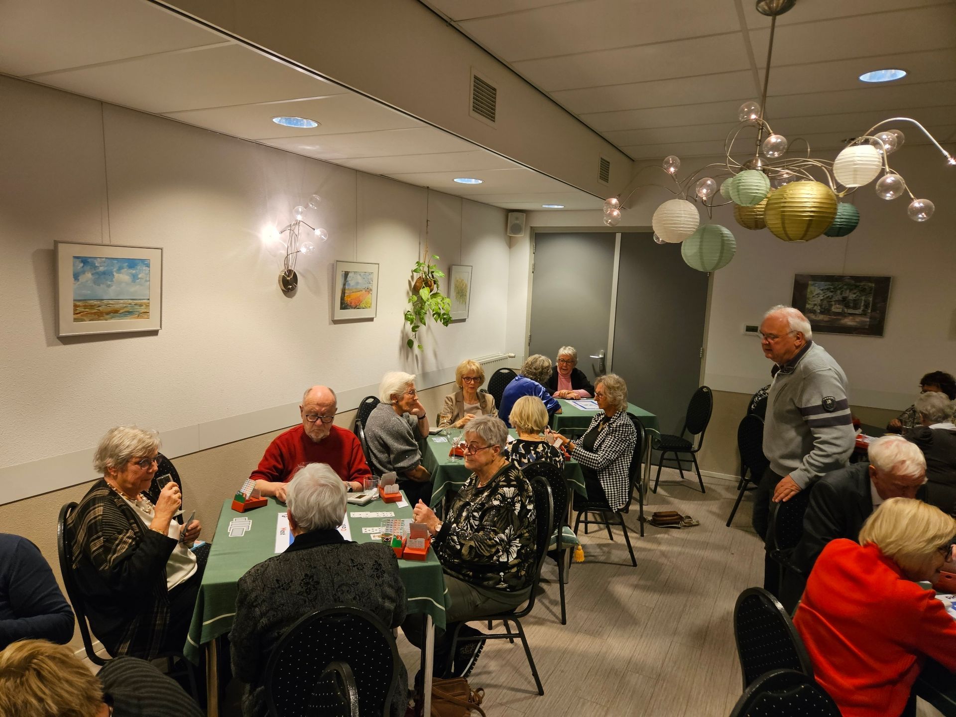 Mensen zitten aan tafels en spelen kaart in een felverlichte ruimte. Sommigen kijken naar de kaarten en praten met elkaar.