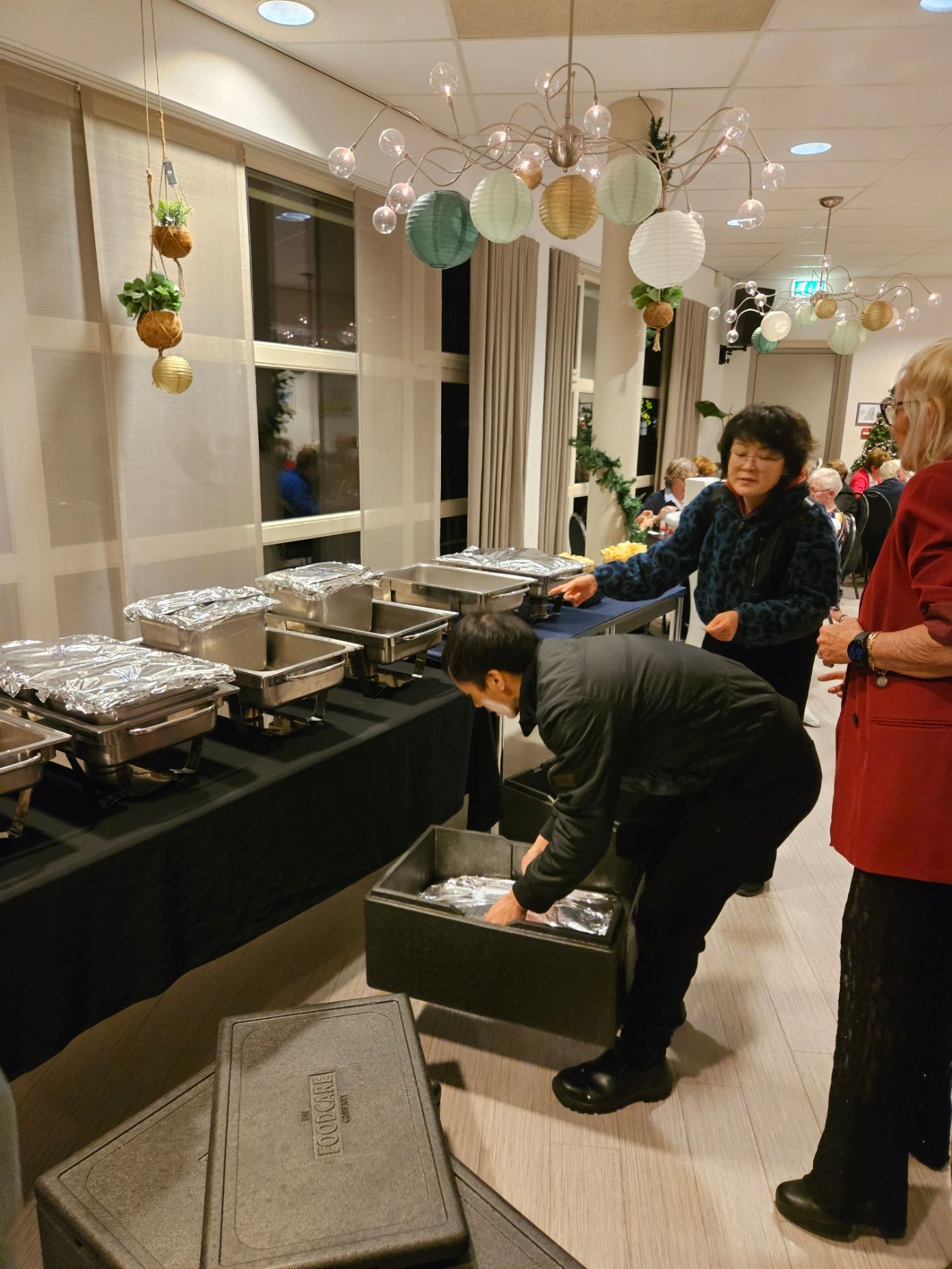 Mensen die eten serveren op een evenement. Buffetopstelling met in folie afgedekte schalen, een zwart tafelkleed en decoratieve verlichting.