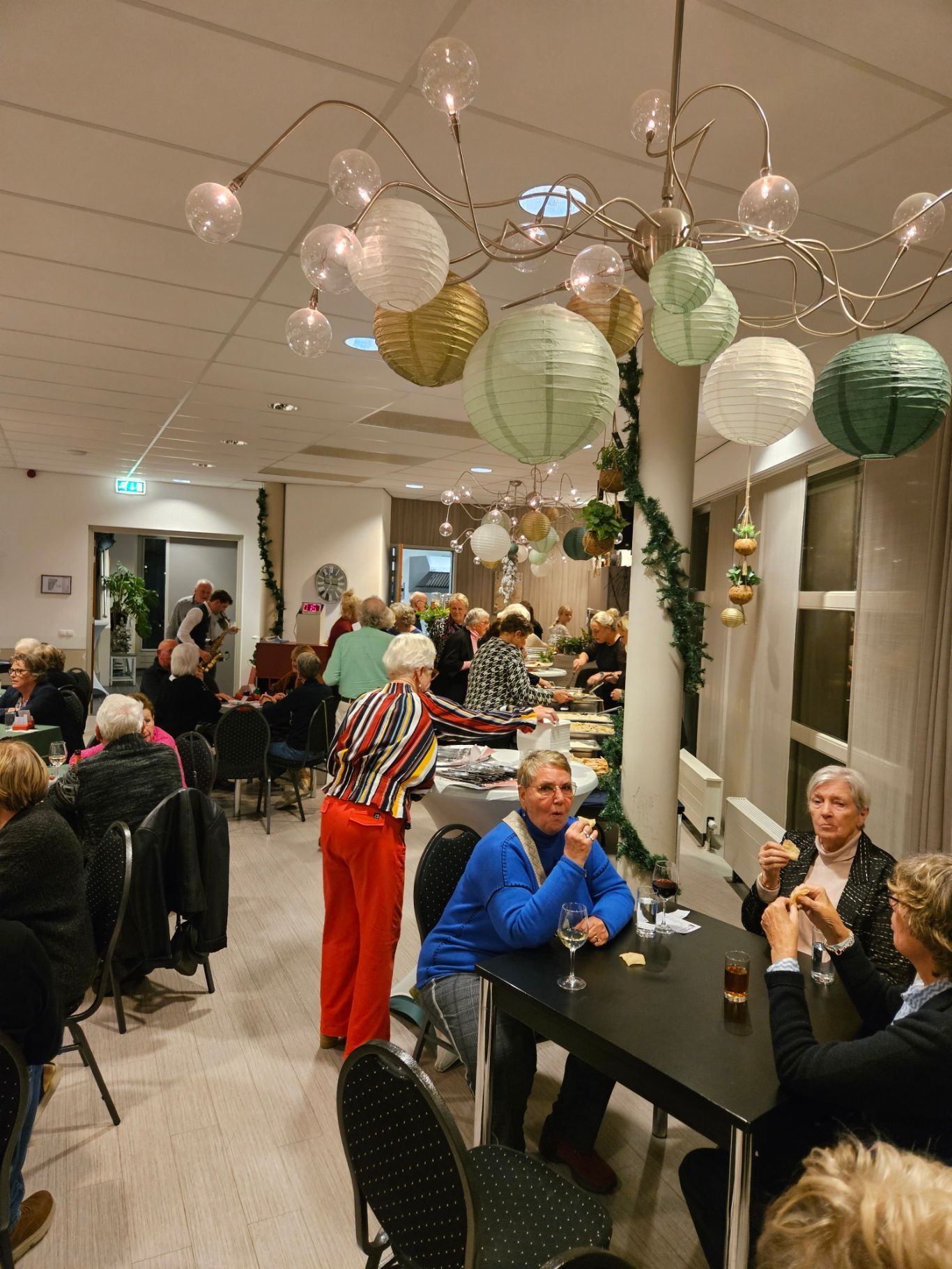 Feestscène: Mensen verzameld in een feestelijk versierde ruimte, met onder andere papieren lantaarns. Sommigen zitten en praten met elkaar.
