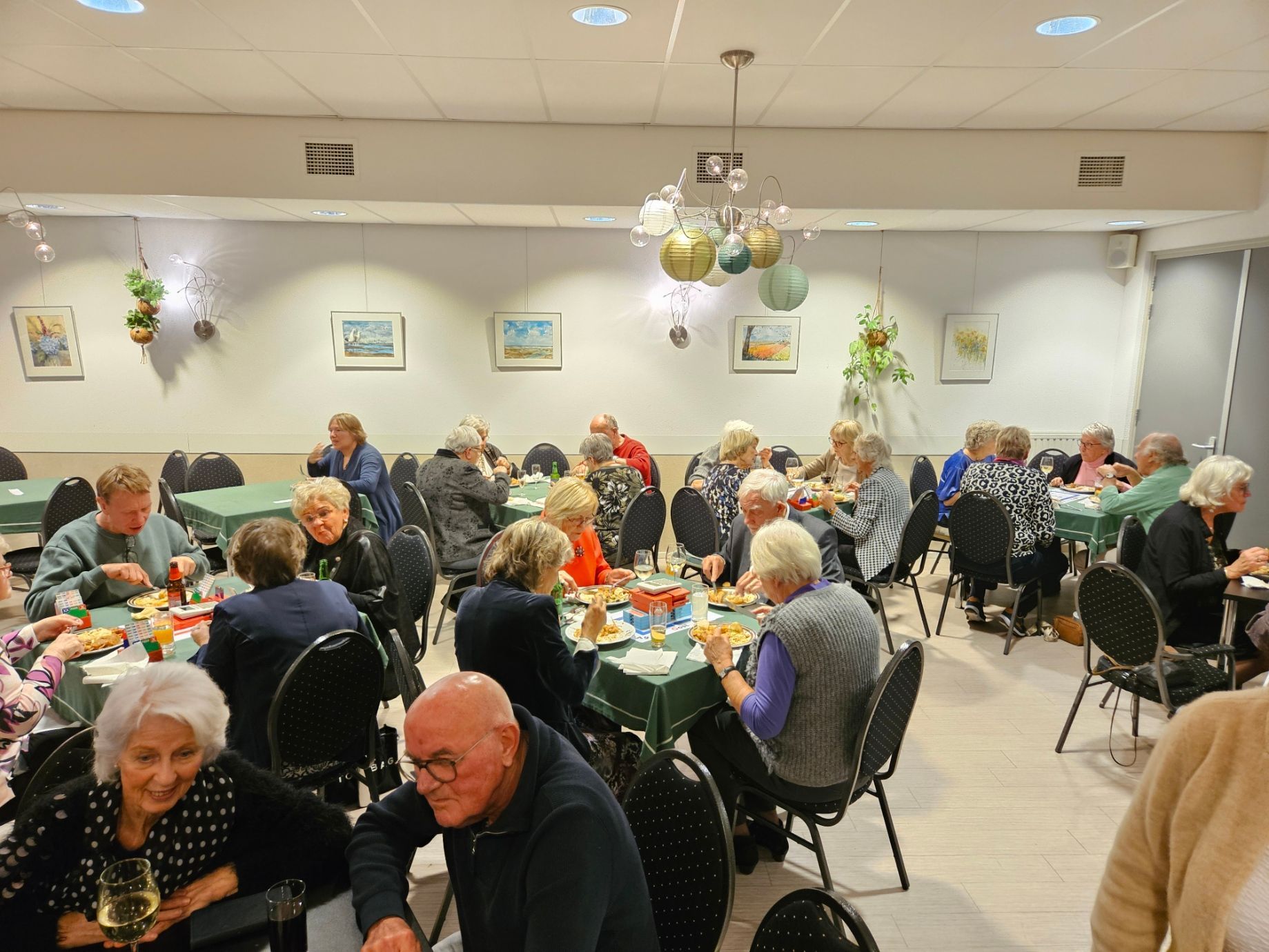 Mensen zitten aan tafels te eten en te praten in een helder verlichte ruimte met kunstwerken aan de muren.