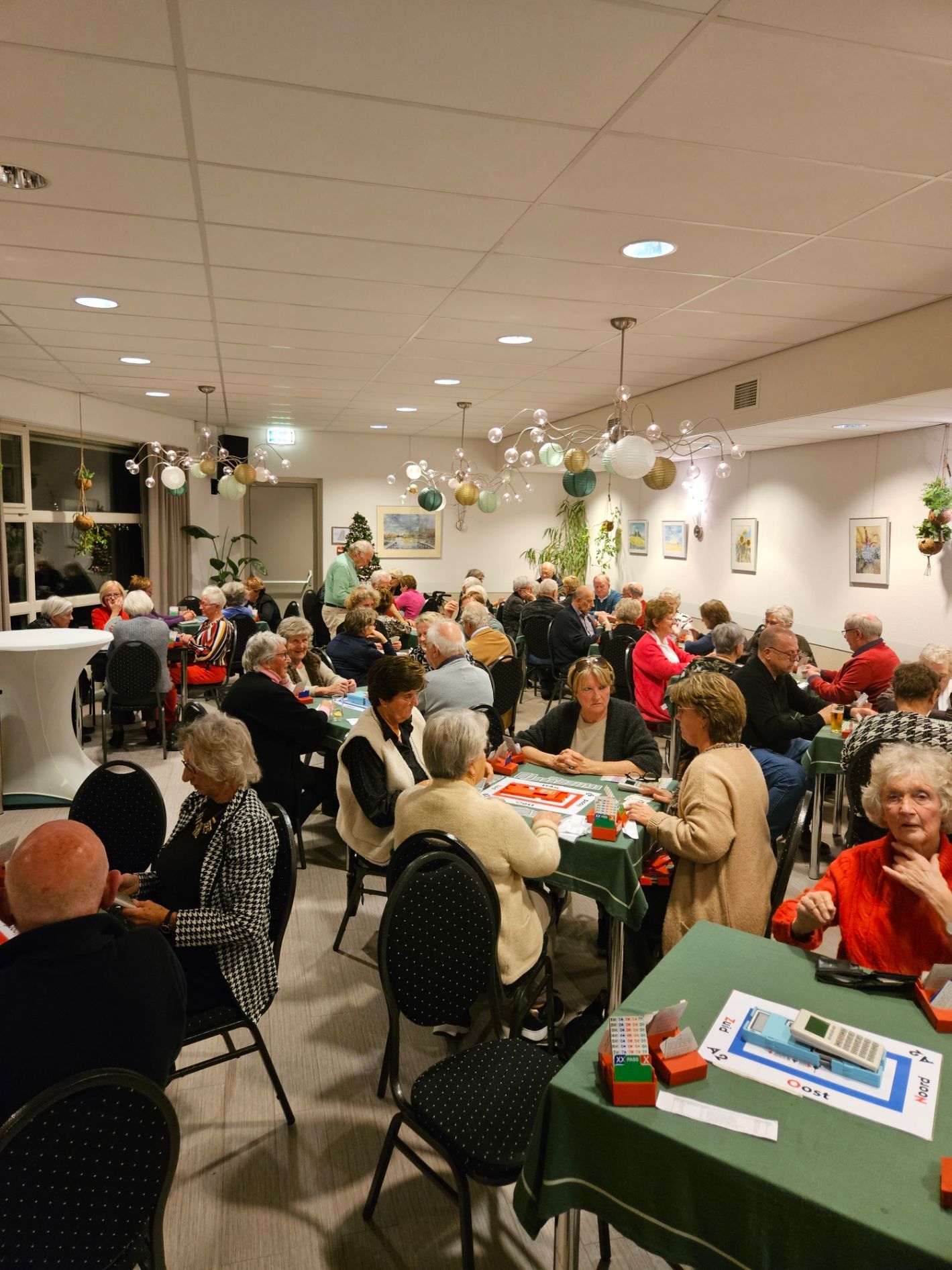 Een grote groep mensen in een goed verlichte ruimte; mensen zitten aan tafels en spelen waarschijnlijk een spel.