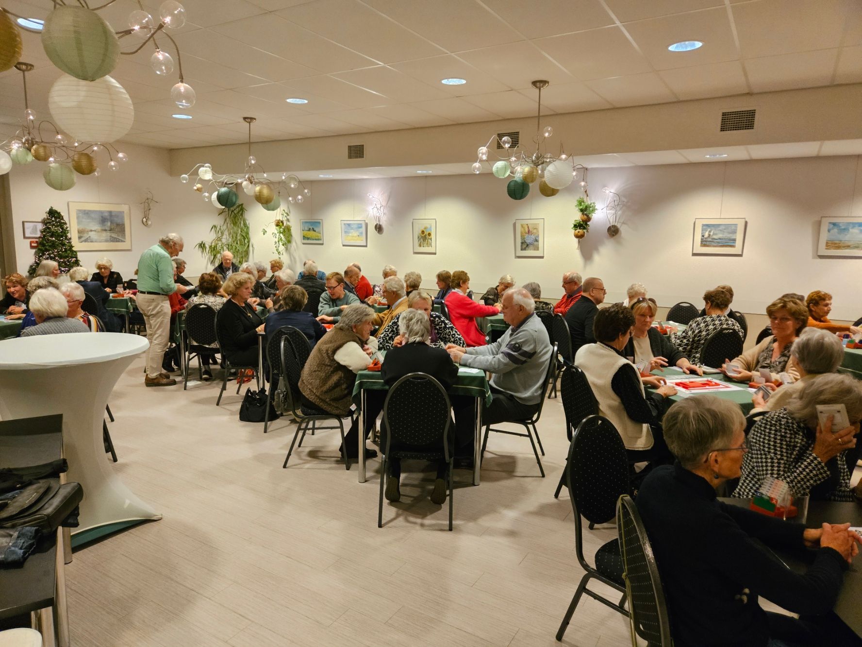 Mensen spelen kaart aan tafels in een helder verlichte gemeenschapsruimte, versierd met hanglampen en kunstwerken.