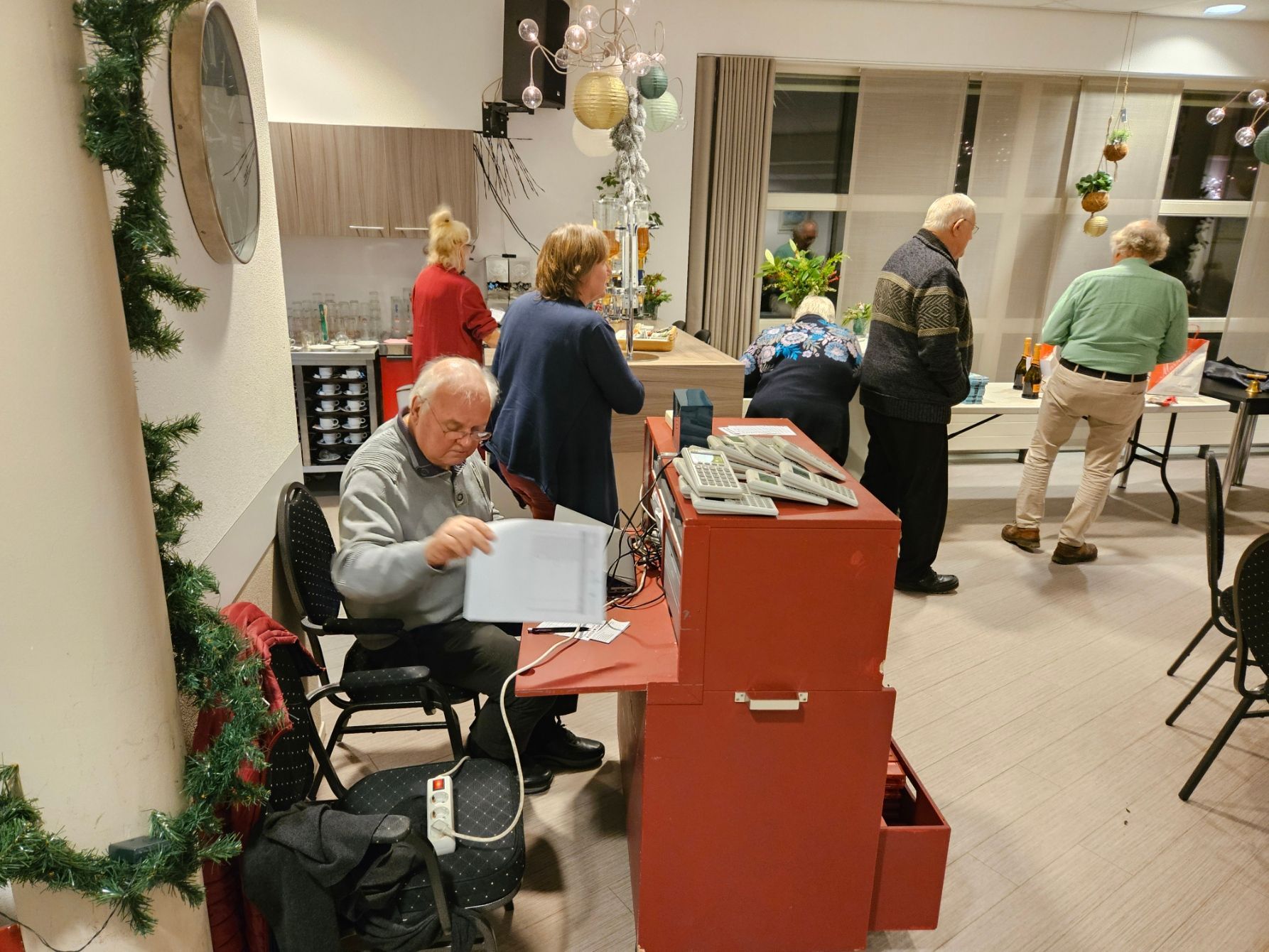 Mensen verzamelen zich in een kamer. Een man zit met papieren in zijn hand. Anderen staan ​​bij een tafel, met op de achtergrond een versierde toonbank.