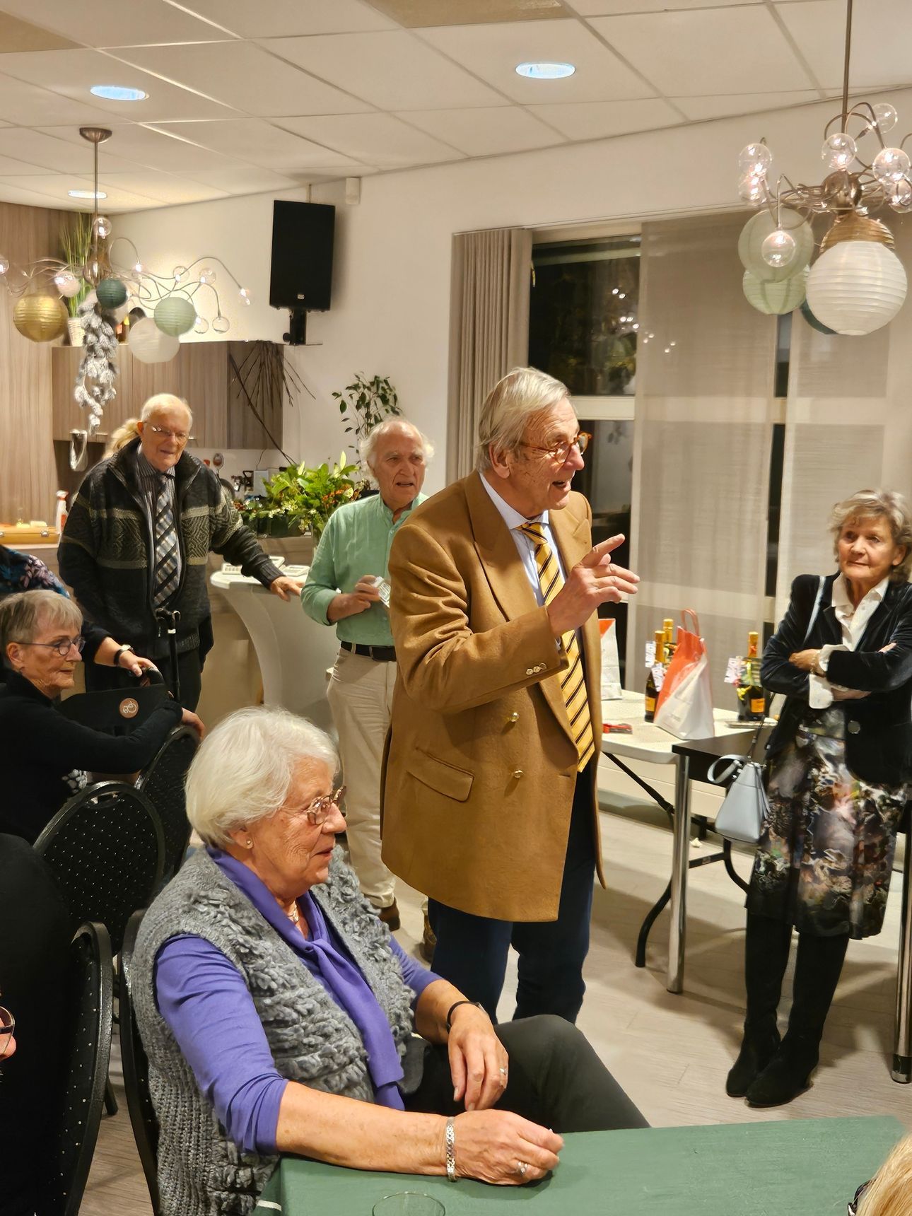 Een man in een bruine jas spreekt een groep toe in een versierde kamer. Anderen luisteren aandachtig, sommigen glimlachen.
