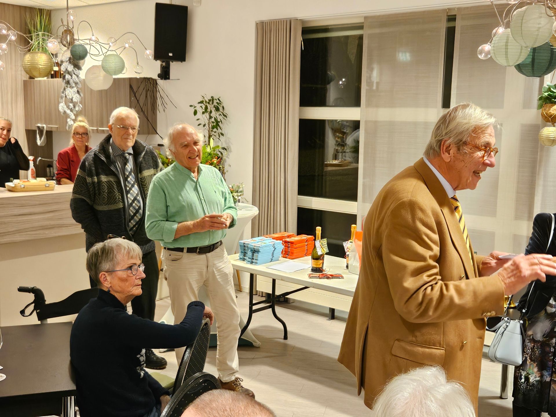 Mensen in een versierde kamer tijdens een bijeenkomst. Een man in een beige jas spreekt; anderen luisteren.