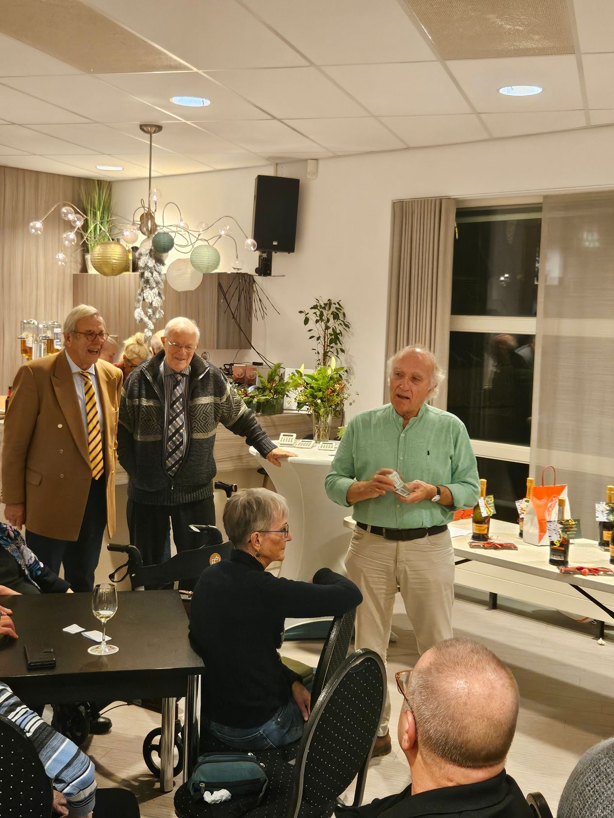 Een man spreekt tijdens een evenement; anderen luisteren. De kamer is versierd en er staat een tafel met flessen.