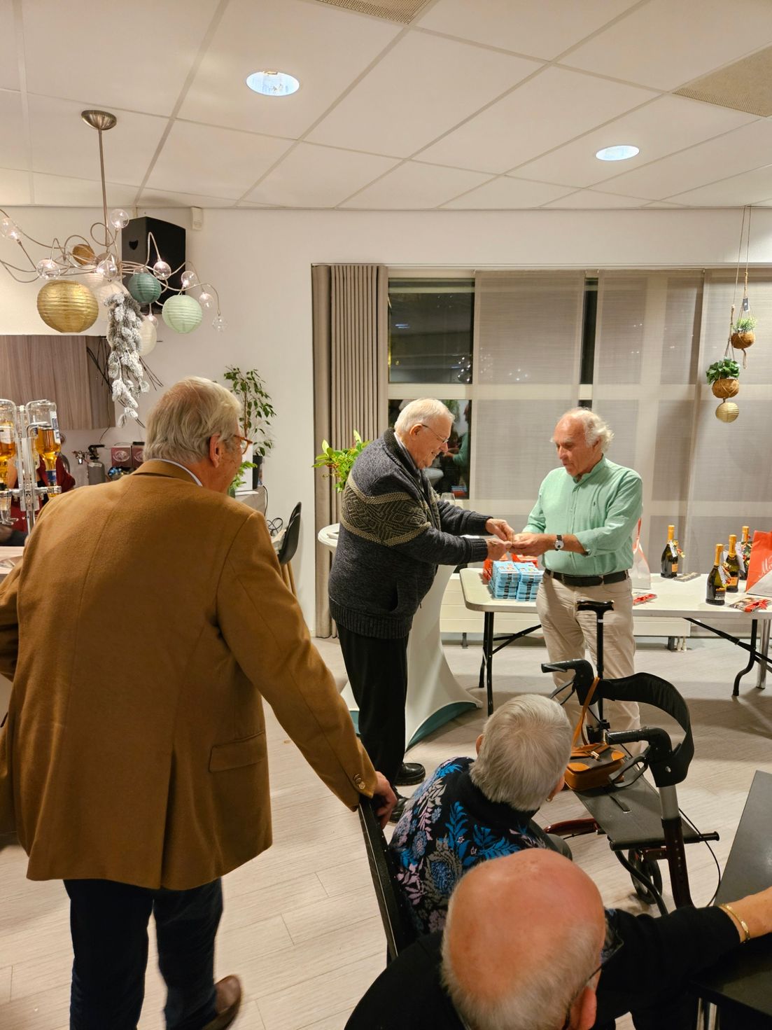 Drie oudere mannen in een kamer, twee schudden elkaar de hand, de derde kijkt toe. Feestelijke versieringen, neutrale achtergrond.