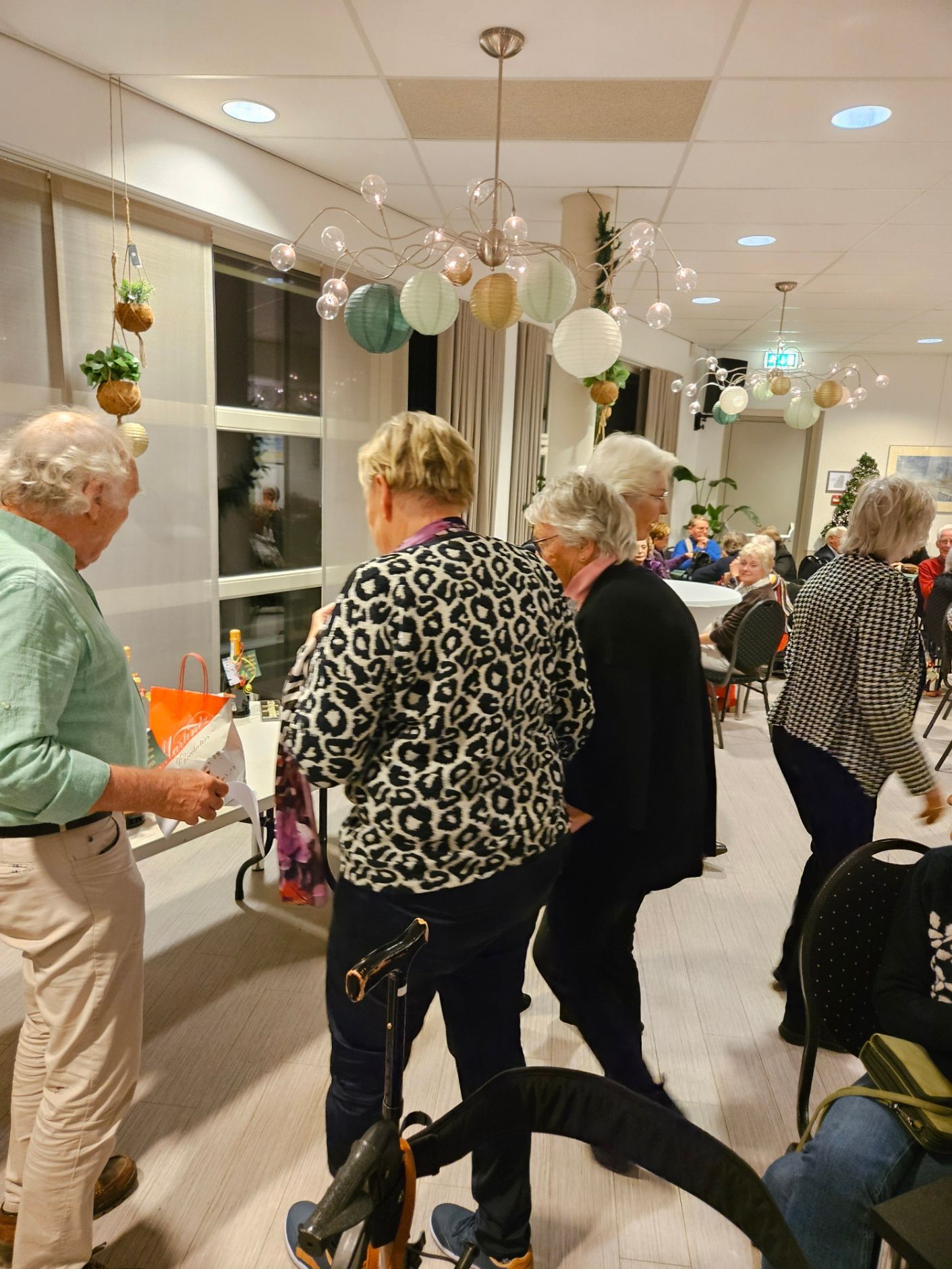 Mensen praten met elkaar op een evenement; boven hen hangt een kroonluchter. Sommigen staan ​​bij een tafel met spullen, anderen lopen eromheen.