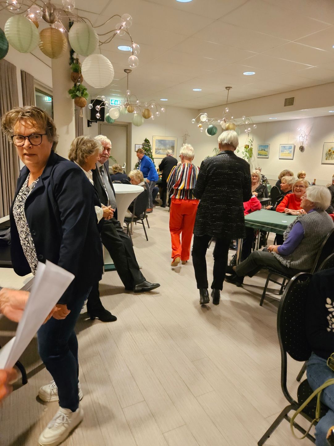 Mensen die gezellig met elkaar praten in een helder verlichte ruimte met witte tafels en stoelen. Sommigen staan, anderen zitten.
