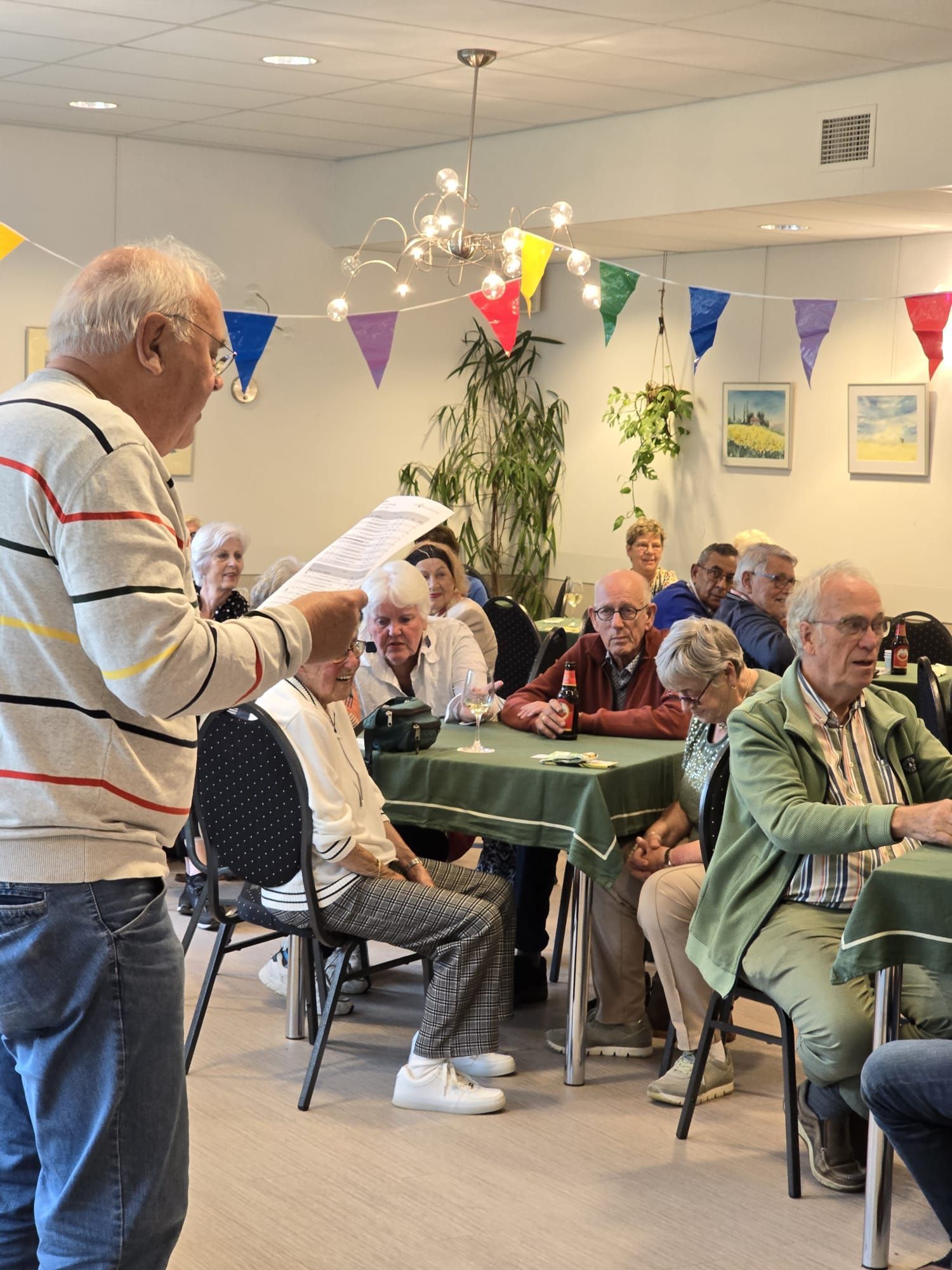 Een man spreekt een groep ouderen toe die aan tafels zitten in een kamer die versierd is met vlaggetjes.