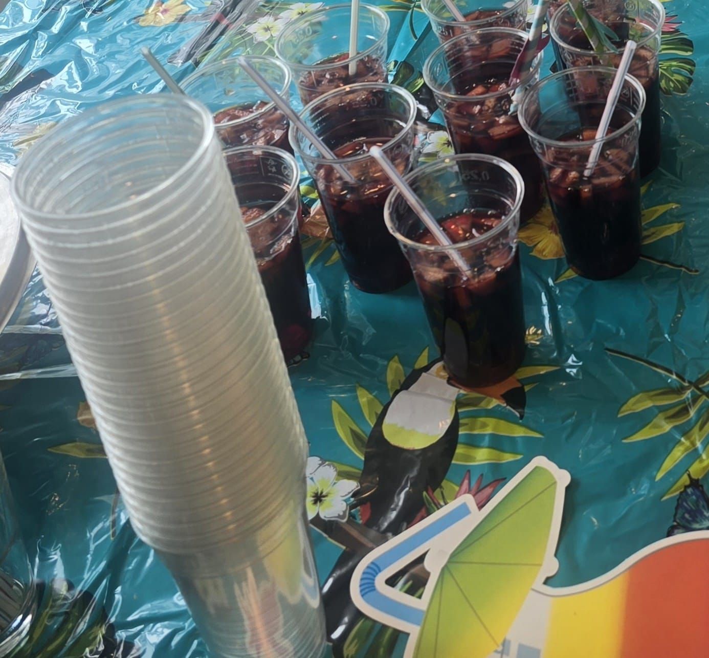 Doorzichtige plastic bekers met donkere drankjes en rietjes, naast een stapel lege bekers op een tafel met een patroon.