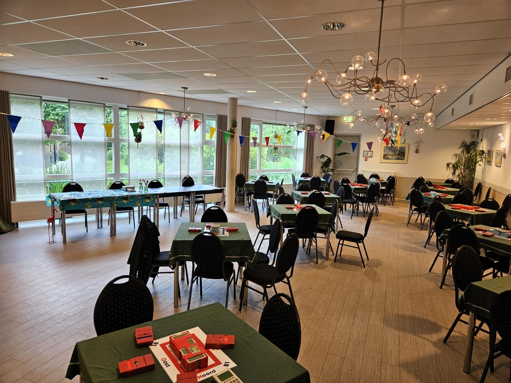 Een grote kamer met tafels en stoelen, klaar voor een kaartspel, versierd met vlaggen en een kroonluchter.
