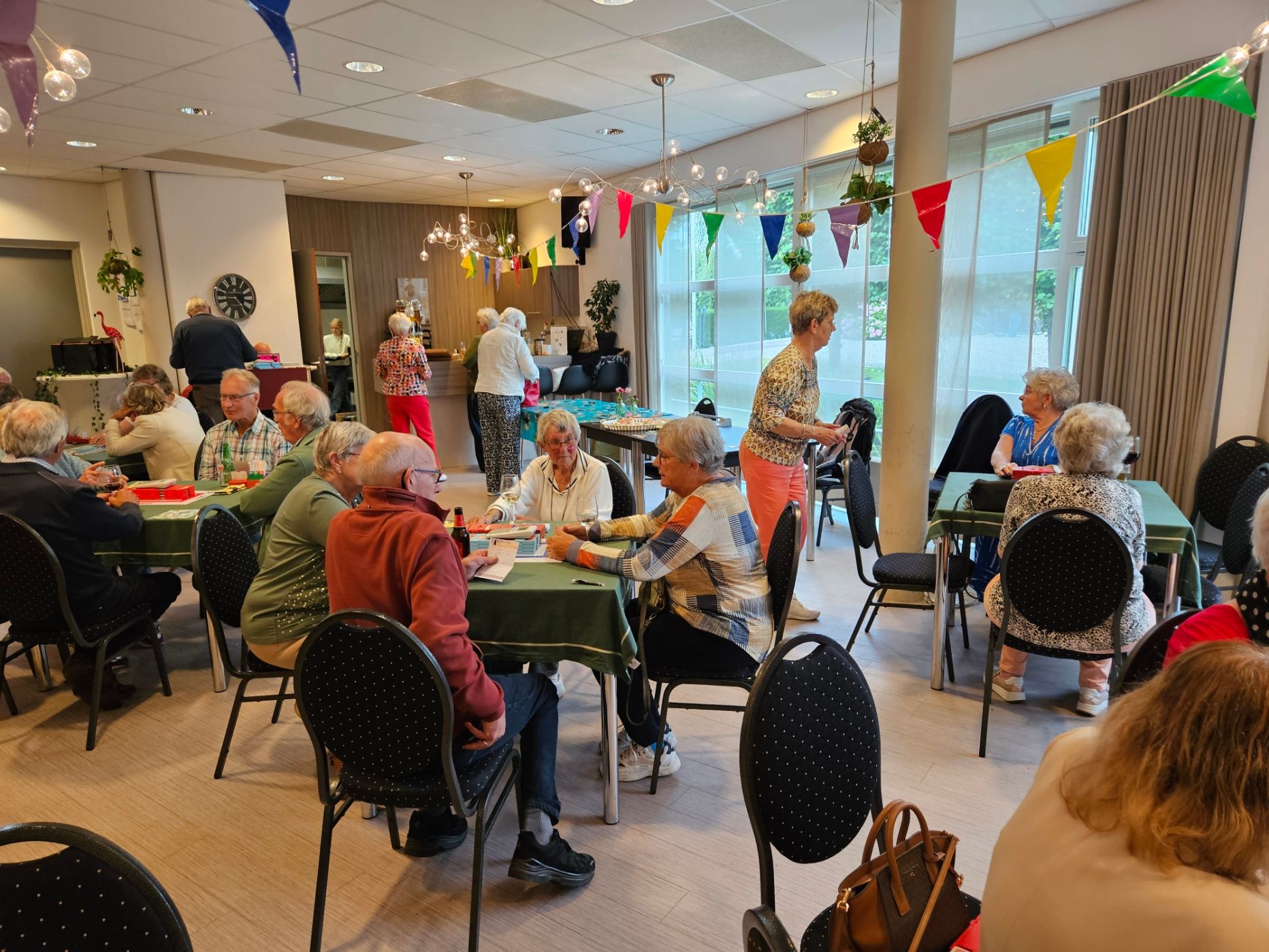 Senioren spelen spelletjes aan tafels in een vrolijk ingerichte ruimte, praten met elkaar en lachen.