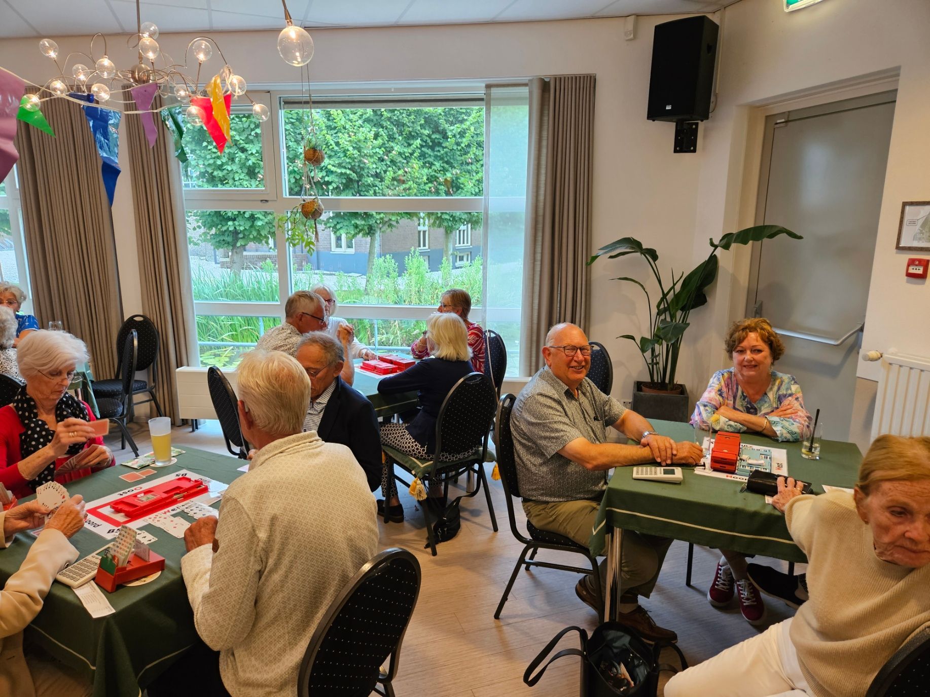 Mensen spelen spelletjes aan tafels in een helder verlichte ruimte met een raam, planten en kleurrijke versieringen.