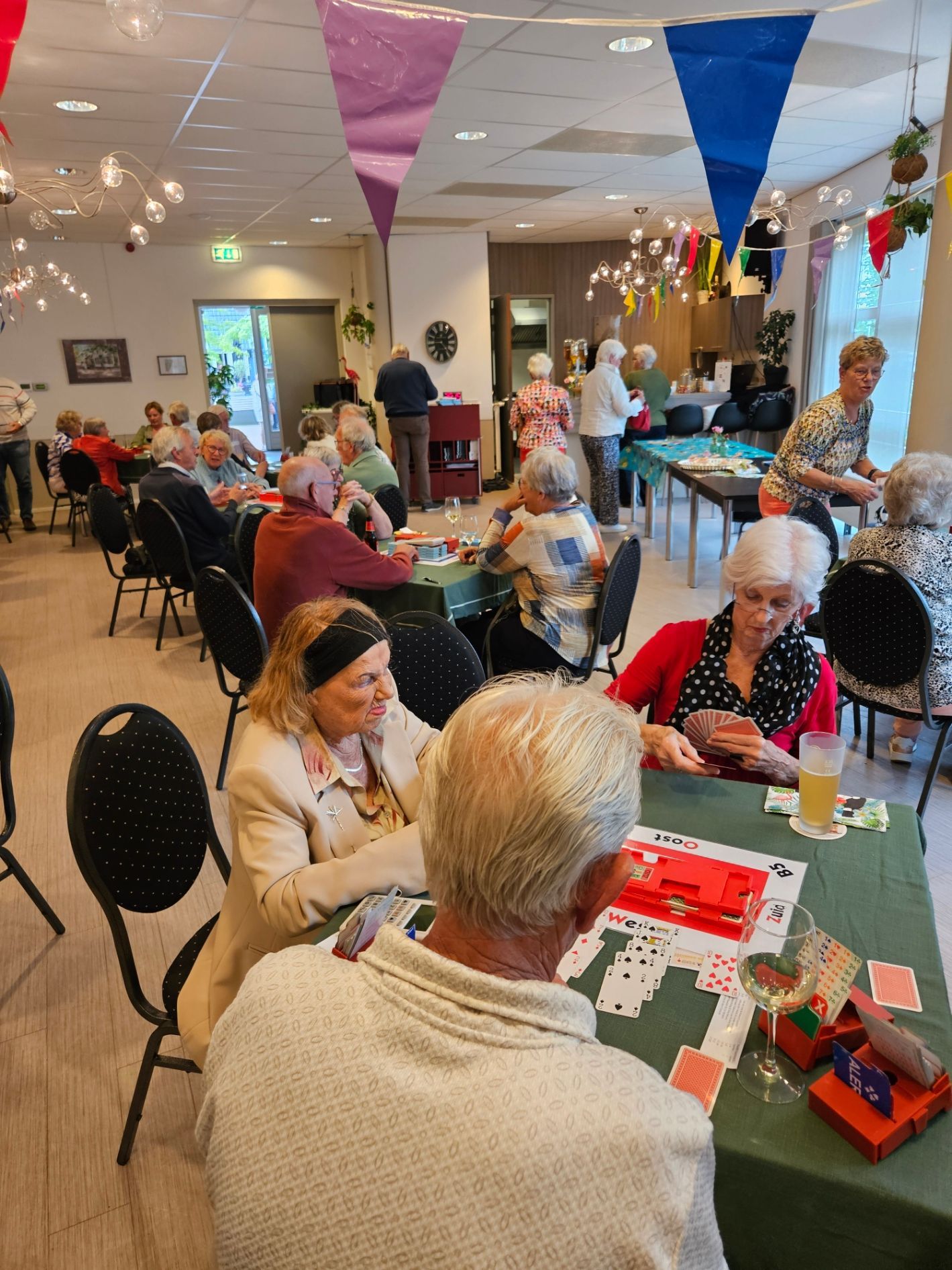 Senioren spelen spelletjes aan tafels in een versierde kamer en genieten van een gezellig samenzijn met kleurrijke slingers boven hun hoofden.