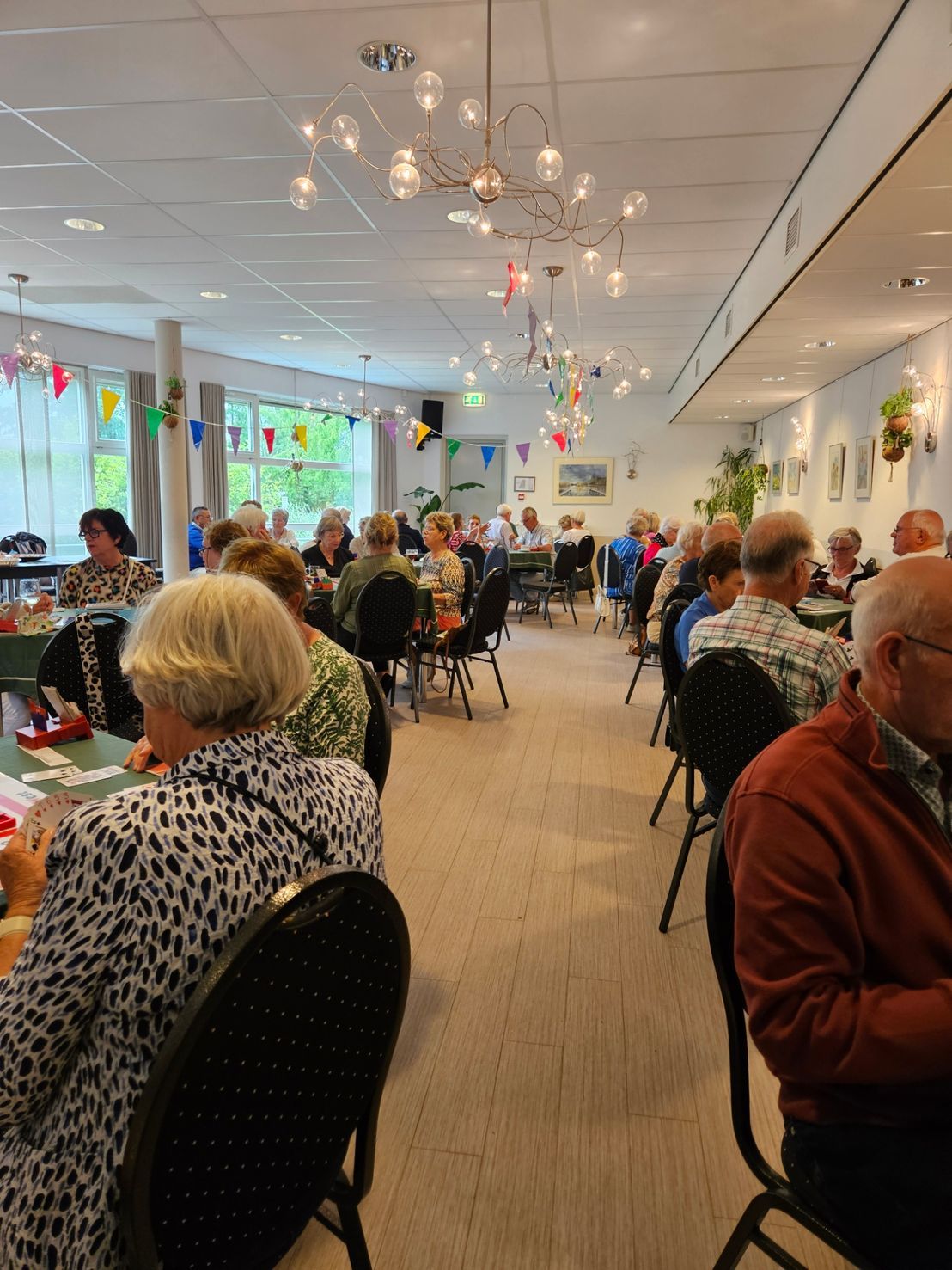Mensen zitten aan tafels in een helder verlichte ruimte. Sommigen spelen kaart, met feestelijke versieringen aan de muur.