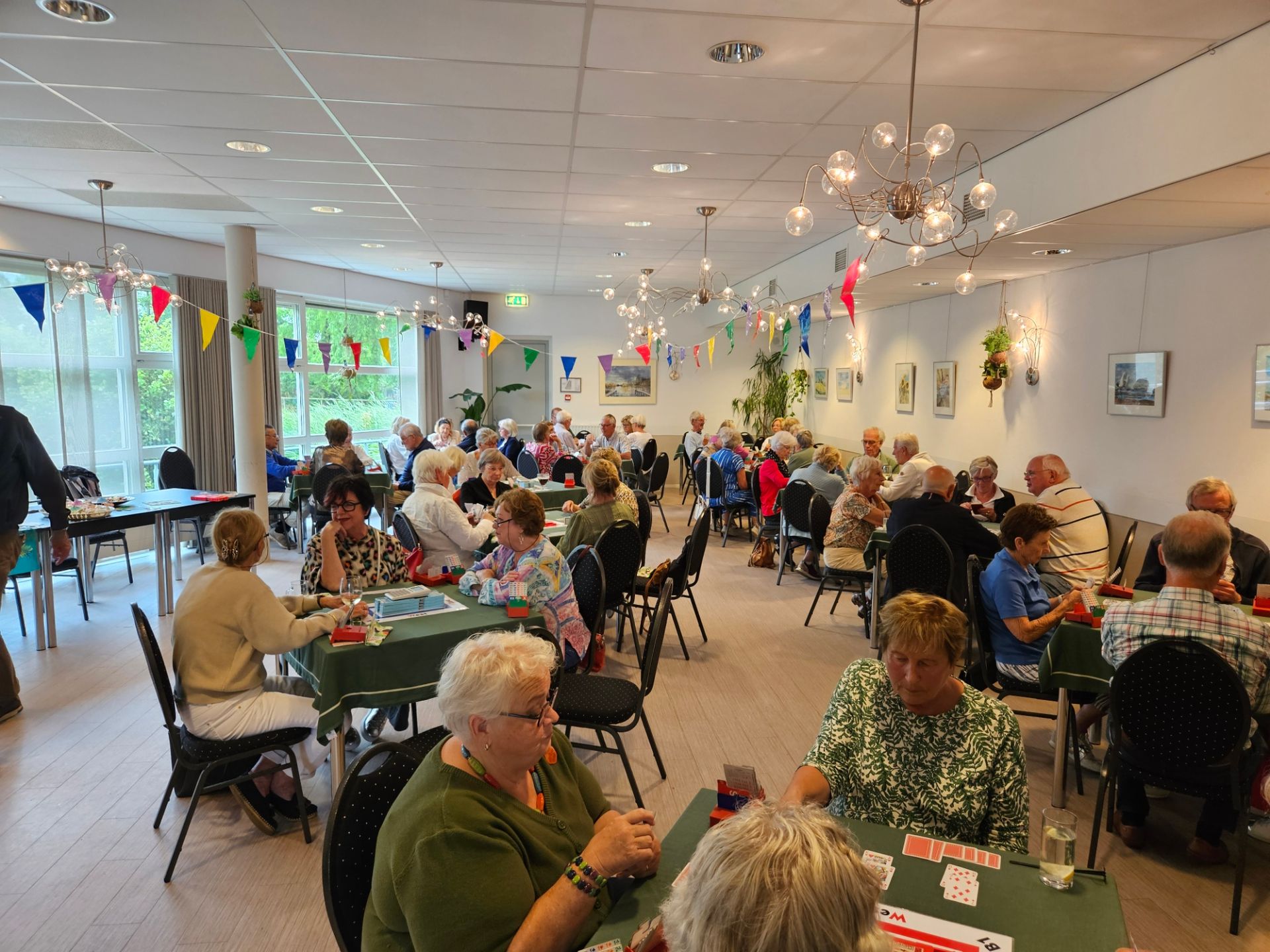 Senioren spelen kaart aan tafels in een helder verlicht gemeenschapscentrum, versierd met vlaggen en hangende lampjes.