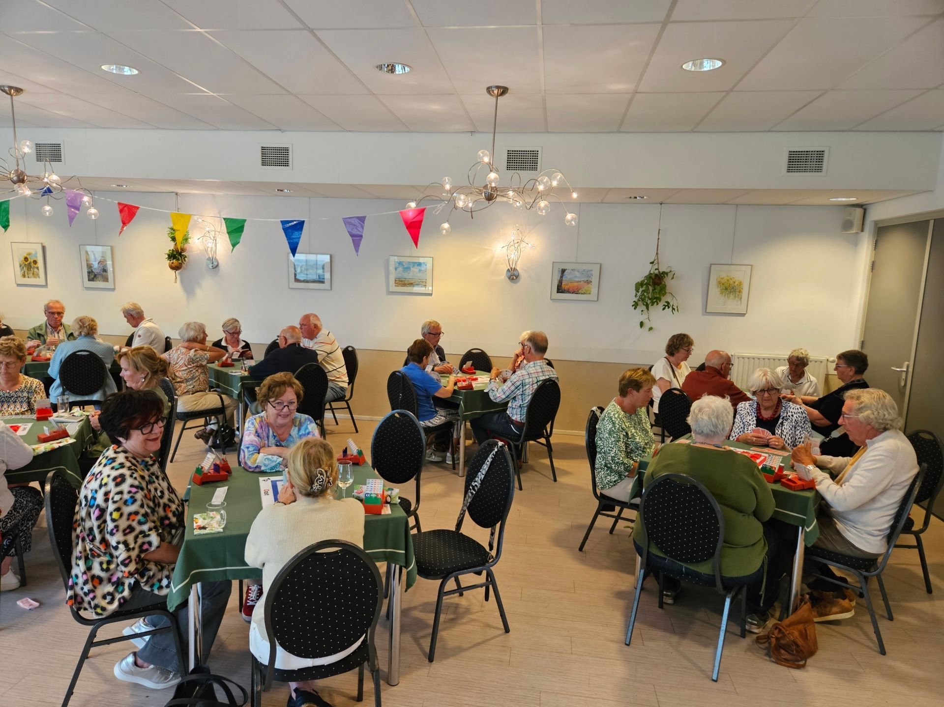 Mensen zitten aan tafels en spelen spelletjes in een gemeenschapscentrum. De tafels zijn gedekt met groene tafelkleden en de muren zijn versierd.