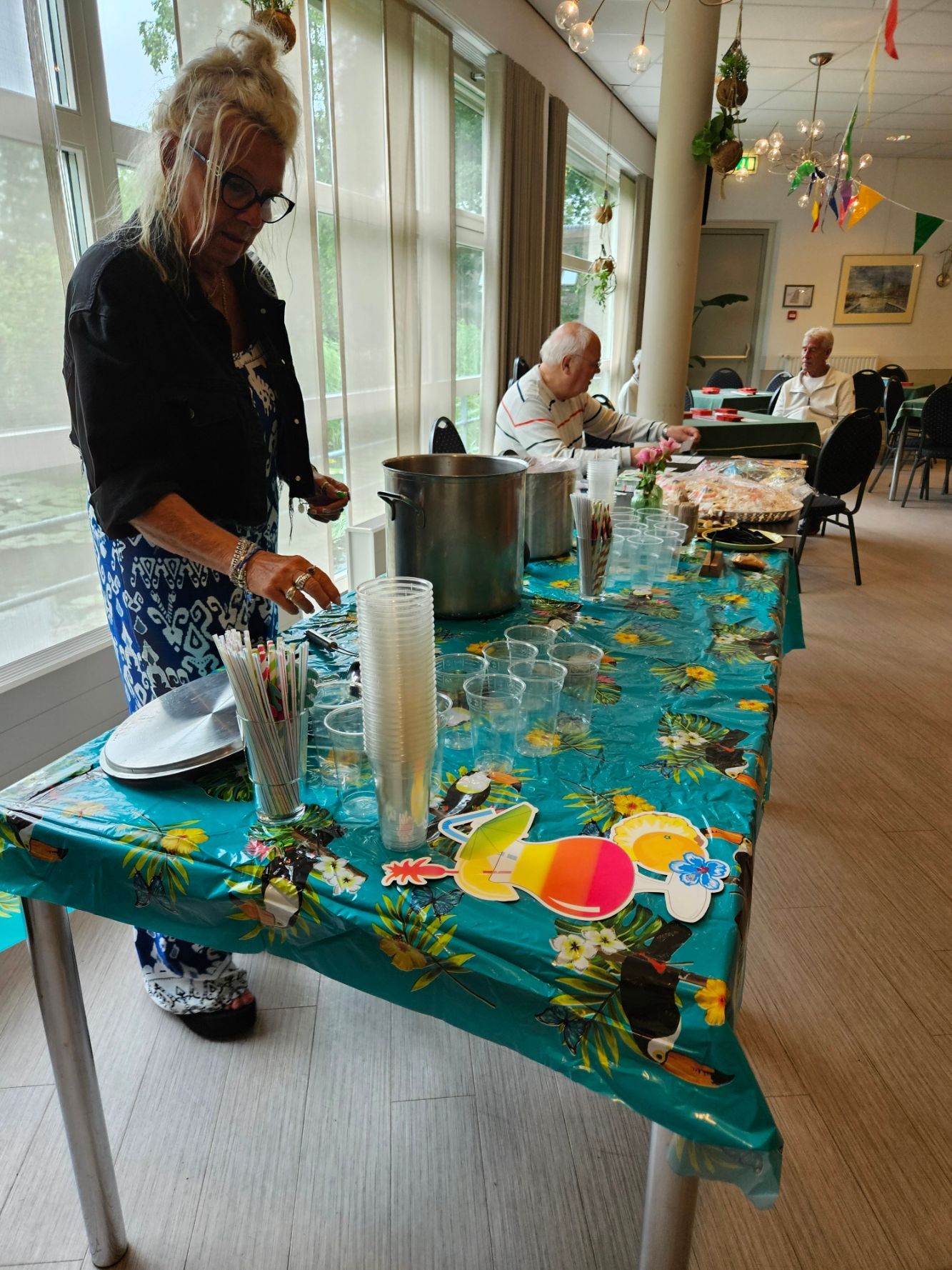 Een vrouw dekt een tafel met glazen en eten in een felverlichte ruimte, met mensen die op de achtergrond zitten.