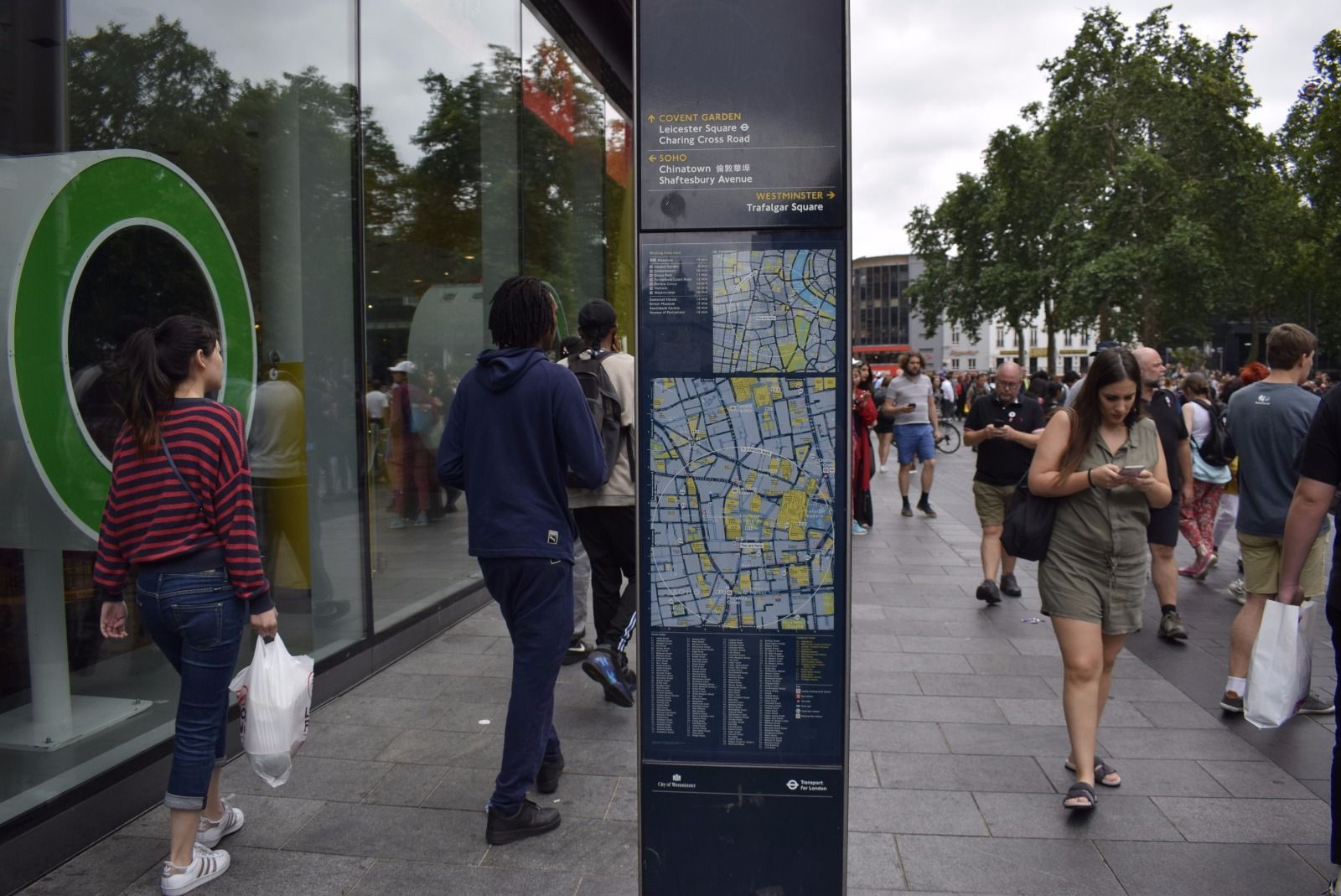 Pedestrians walk along a sidewalk past a glass building and a map kiosk in a busy, urban area.