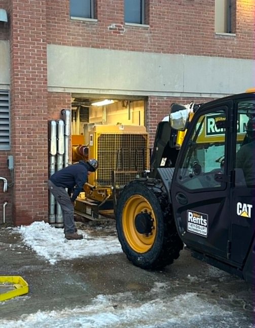 Worker loads equipment into a CAT forklift beside a brick building in a snowy loading area.