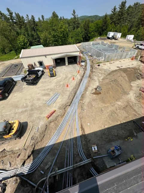 Road construction site with excavators, utility conduits, and a small building near a wooded area